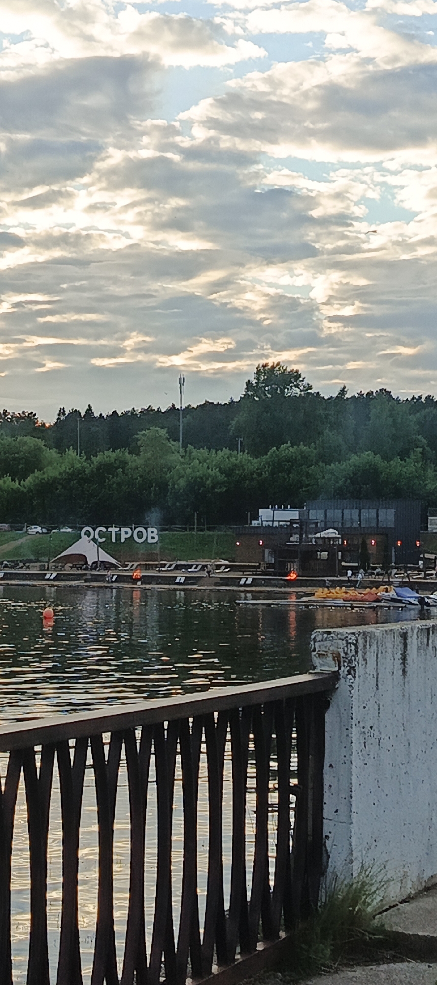 This media file captures a scenic view of a waterfront area with a large sign spelling out "ОСТРОВ" (Ostrov, meaning Island) in white block letters. The scene appears to be set in a recreational area by a body of water, possibly a lake or river, with a treeline in the background.
In the foreground, a dark metal railing with an artistic, curved design separates the viewer from the water. The water itself reflects the sky and surrounding lights, creating shimmering patterns. On the water, a bright orange buoy is visible.
Beyond the water, a waterfront promenade is lined with lounge chairs and a large, light-colored tent or canopy structure, suggesting a place for relaxation and outdoor activities. There are also what appear to be pedal boats and a small motorboat docked or on display. A dark, building with illuminated windows is situated on the waterfront, with lights reflecting in the water. Small fires or heat lamps are also visible on the dock area, adding a warm glow to the scene.
The background features a dense forest, with a single telecommunication tower rising above the trees. The sky is filled with dramatic clouds, illuminated by the soft, warm light of either sunrise or sunset, indicating a late evening or early morning. The weather appears partly cloudy with some breaks of sunlight.
There are no distinct individuals clearly visible in the frame, though some small figures might be present near the buildings or on the docks. The overall mood of the image is peaceful and serene, highlighting the natural beauty of the location and the leisure amenities offered. The visible text "ОСТРОВ" is a key identifier of the location.