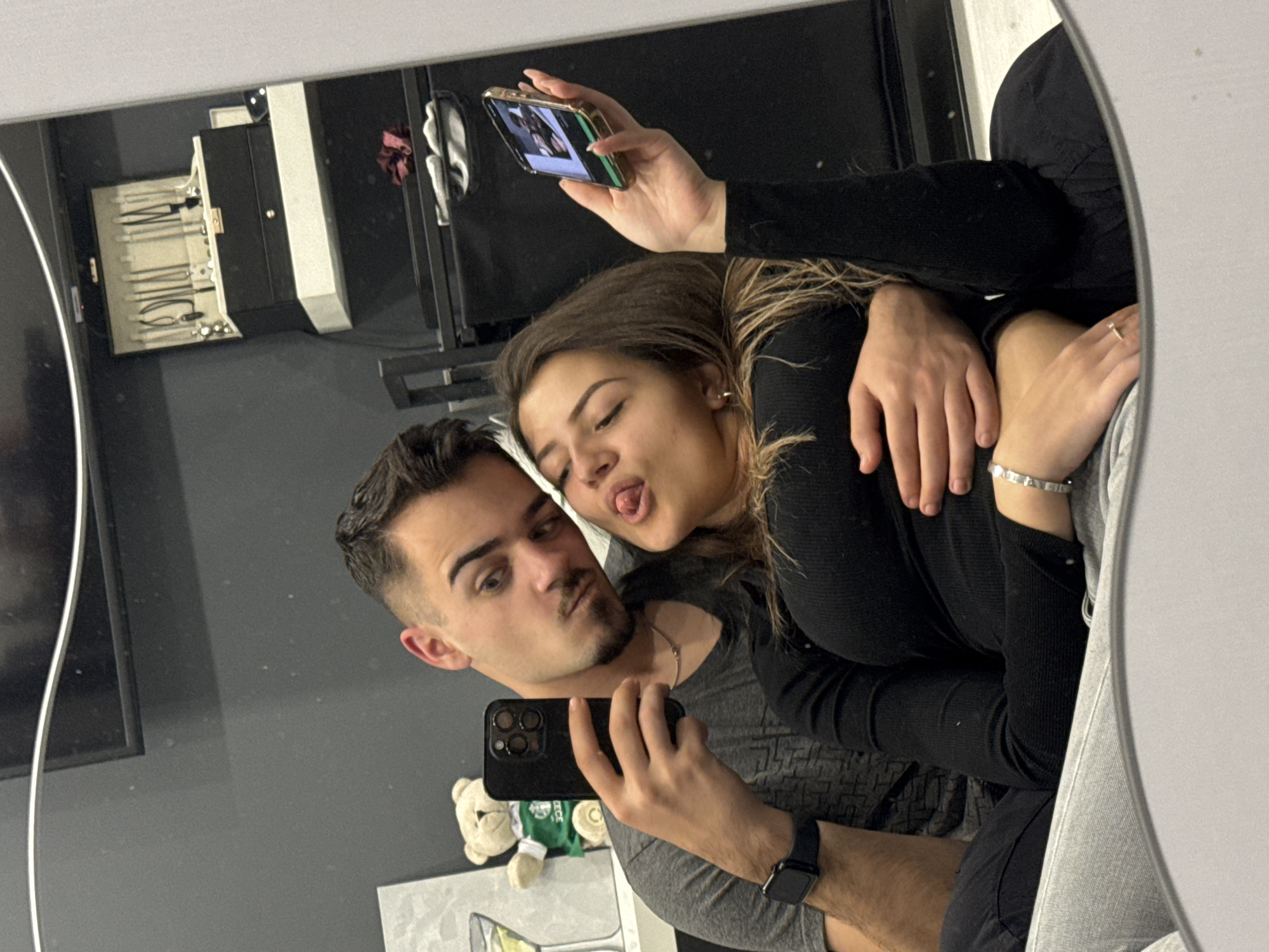 A young couple is taking a selfie in a mirror. The man is holding a smartphone with a green case, wearing a watch on his left wrist, a silver necklace, and a gray t-shirt. The woman is sticking her tongue out and has her arm around the man's shoulder. She is wearing a black long-sleeved shirt and silver jewelry on her wrist. The image is a reflection, and the background includes a black cabinet with jewelry visible in the reflection, a teddy bear on a shelf, and a white surface. The location cannot be determined without more information.
