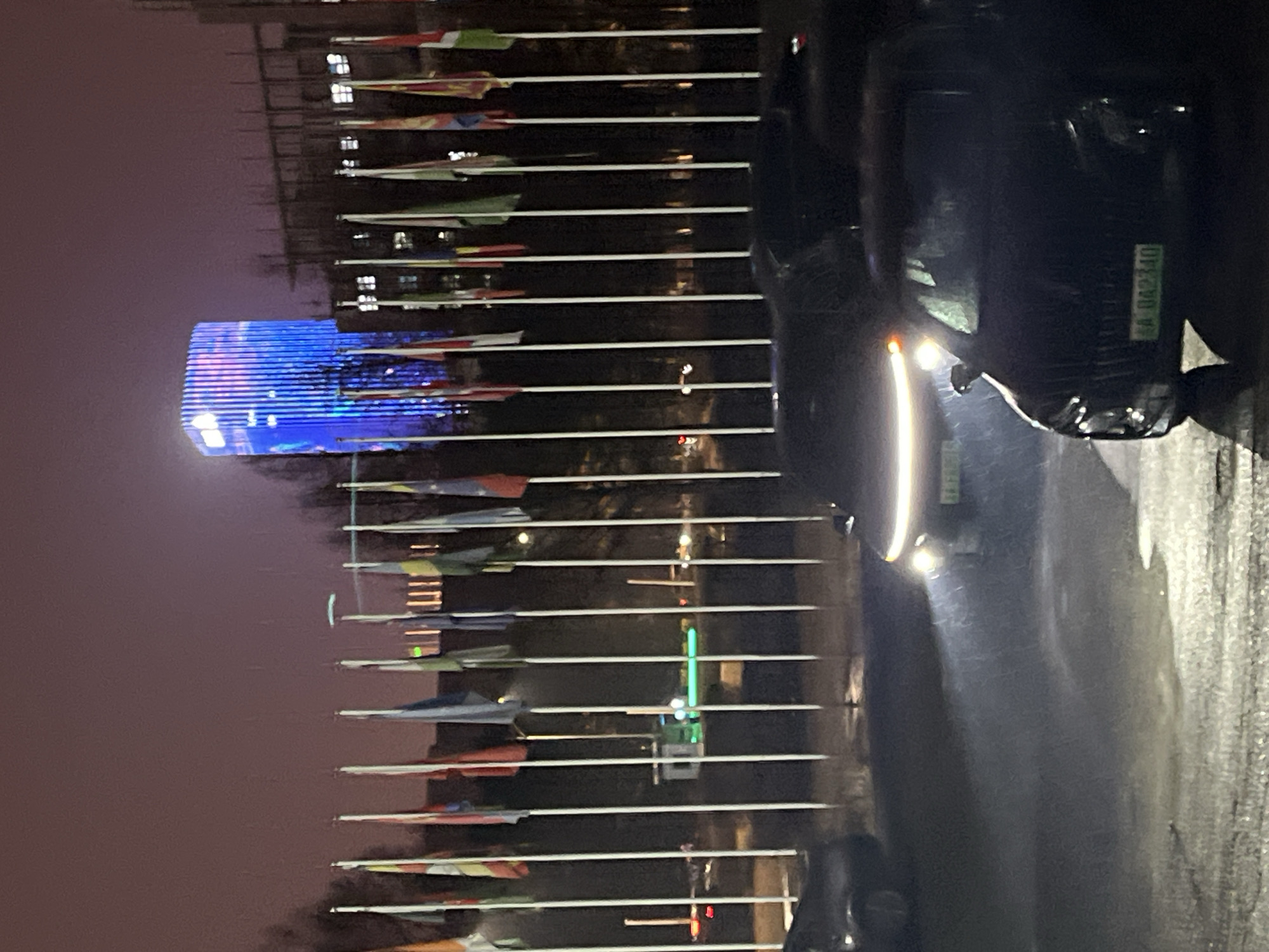 The image shows a night scene in Changchun City, China. A building illuminated with blue lights is visible on the left. A row of flag poles extends towards the right. In the foreground, a dark car is parked on a wet street, its headlights reflecting off the pavement. The license plate of the car reads "吉A 042340." The sky is overcast.