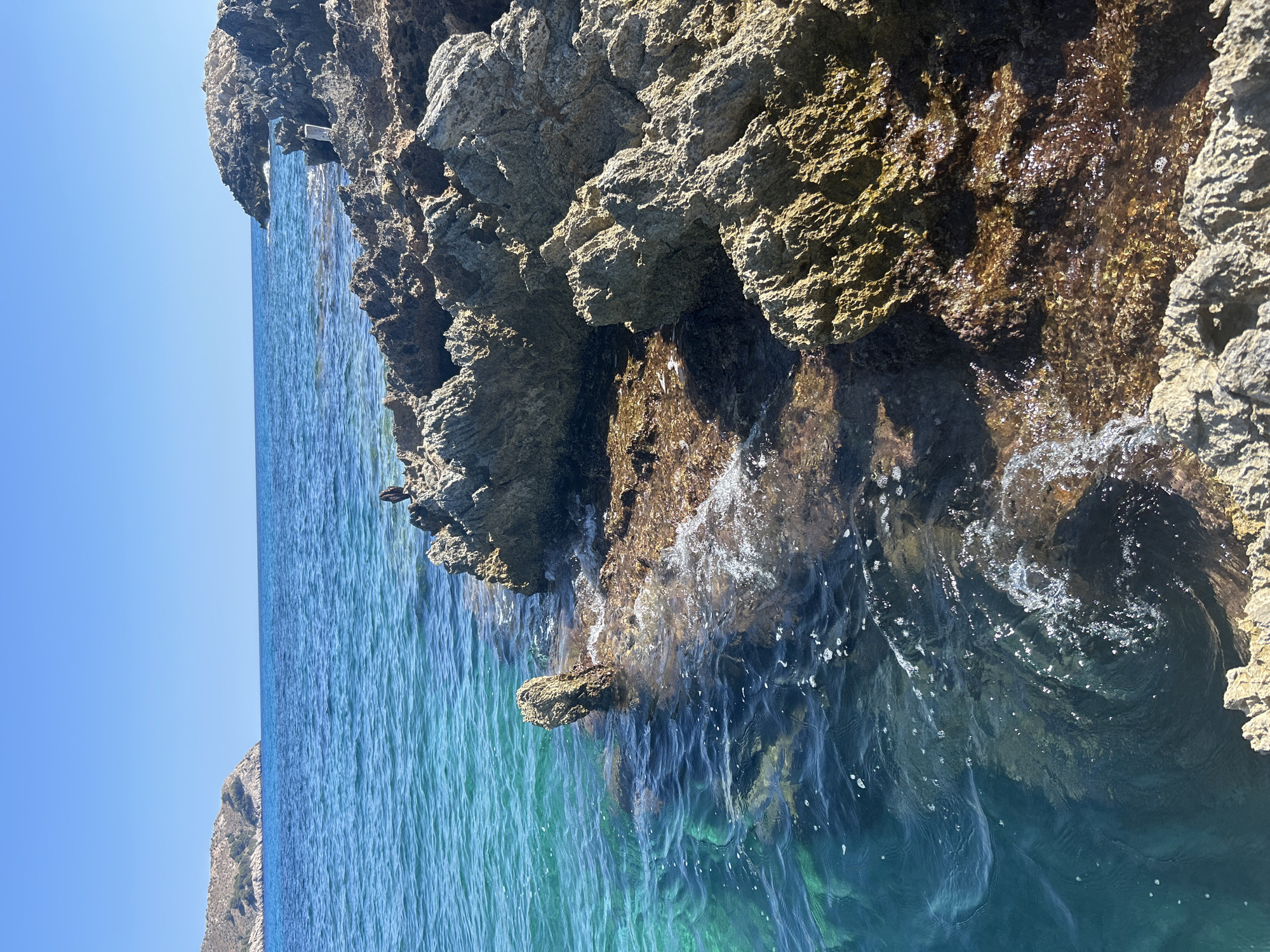 The image depicts a rocky coastline under a clear blue sky. The water is a vibrant turquoise, transitioning to deeper blues further out. Waves gently lap against the jagged, tan and brown rocks that form the shore. In the background, a distant island with a green, hilly landscape is visible. The scene appears to be uninhabited, with no people or animals present. The lighting suggests a bright, sunny day.