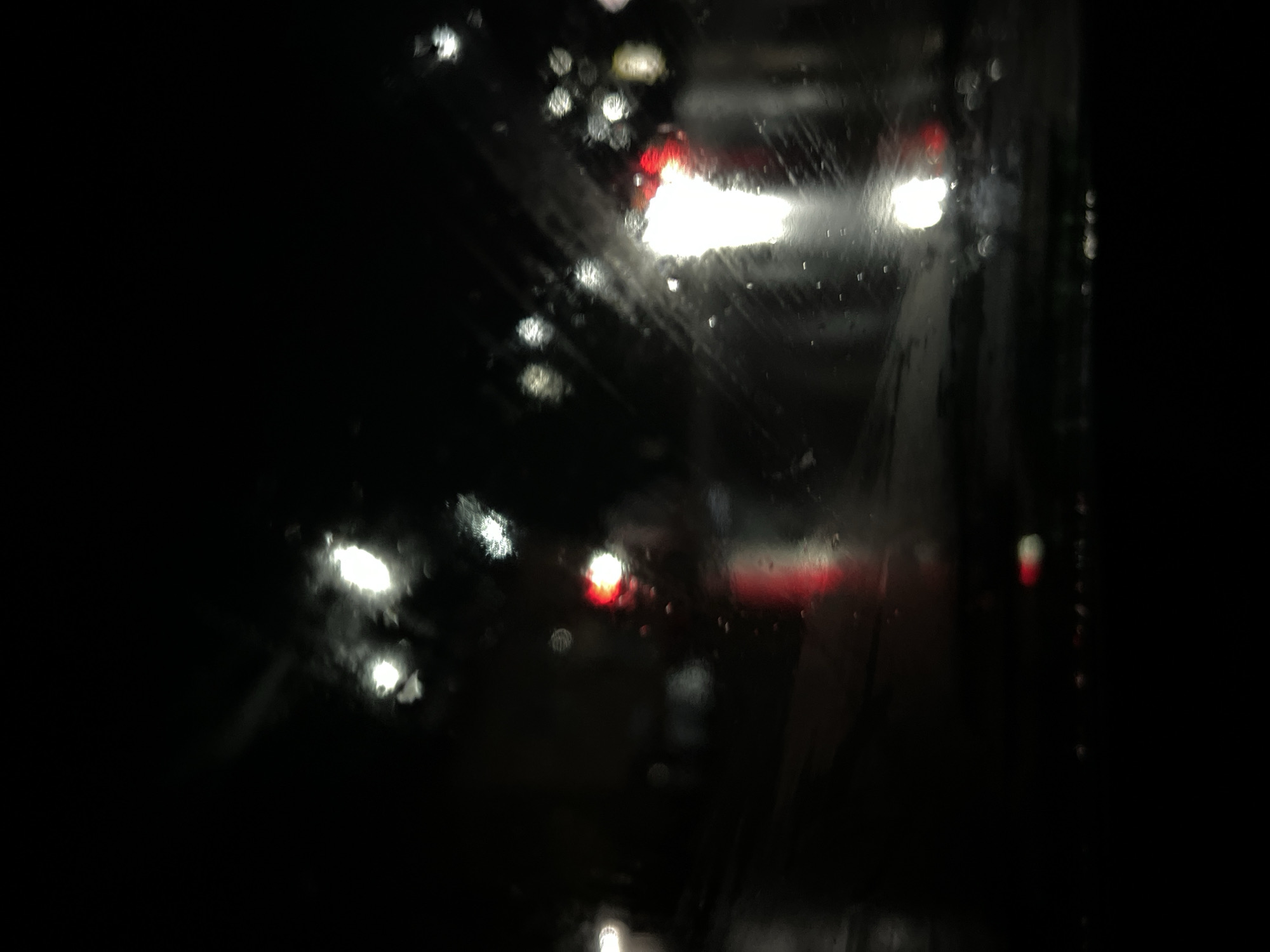 This is a low-light, out-of-focus photograph of traffic at night, possibly taken from inside a vehicle. The scene is dark, with the primary visual elements being blurred lights. There are several bright, white or yellowish lights that appear to be headlights or streetlights, and some red lights that could be taillights or traffic signals. The surface on which the lights are reflected appears wet, suggesting it is raining or the ground is damp, which is further indicated by water droplets visible on the foreground, likely on a window. The overall impression is that of a city street at night during inclement weather. There are no discernible people or specific landmarks that would indicate a precise location within Belogorsk, Russia.