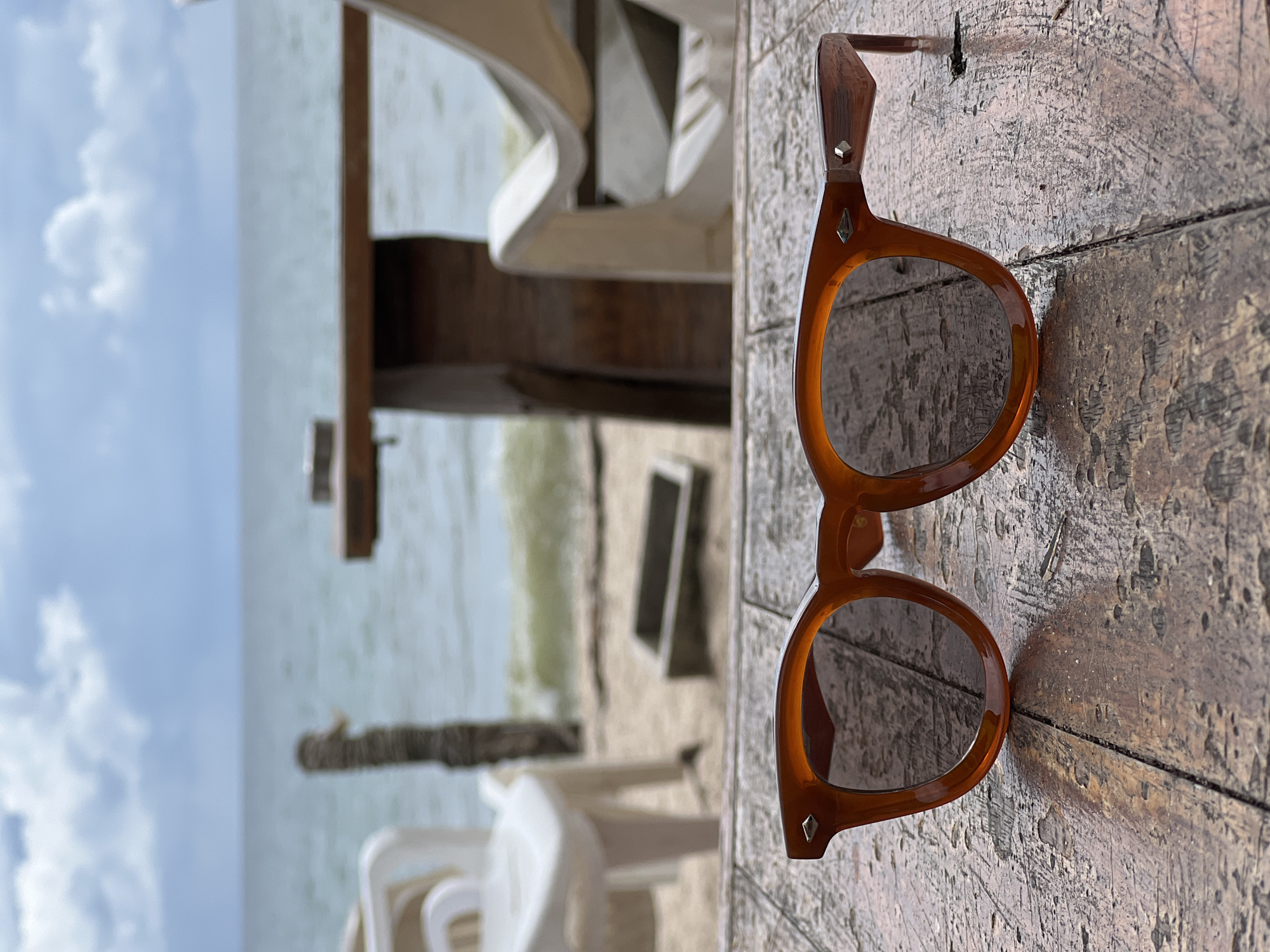 A pair of amber-colored sunglasses with dark lenses rests on a weathered wooden table. The table's surface is rough and shows signs of age, with distinct wood grain and peeling paint. In the background, the ocean is visible, with a slightly overcast sky above. The water is a muted blue-green, and gentle waves are breaking on a sandy beach. A few white plastic chairs and a wooden table are blurred in the mid-ground, suggesting an outdoor seating area near the beach. The overall scene evokes a relaxed, casual atmosphere.