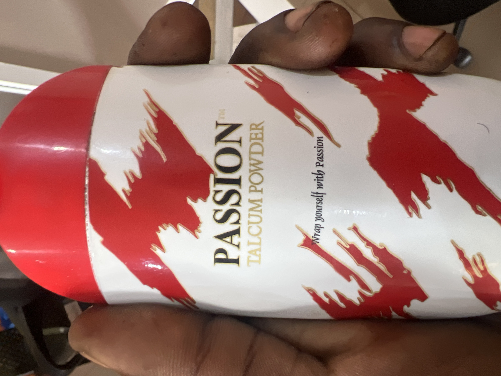 The image shows a hand holding a cylindrical container of "PASSION TALCUM POWDER". The container is white with red abstract designs and black and gold text. The lid of the container is red. The hand appears to be a person of African descent. The background is blurred, but appears to be indoors with some furniture. There is no visible indication of the city of Jalingo, Nigeria in the image.