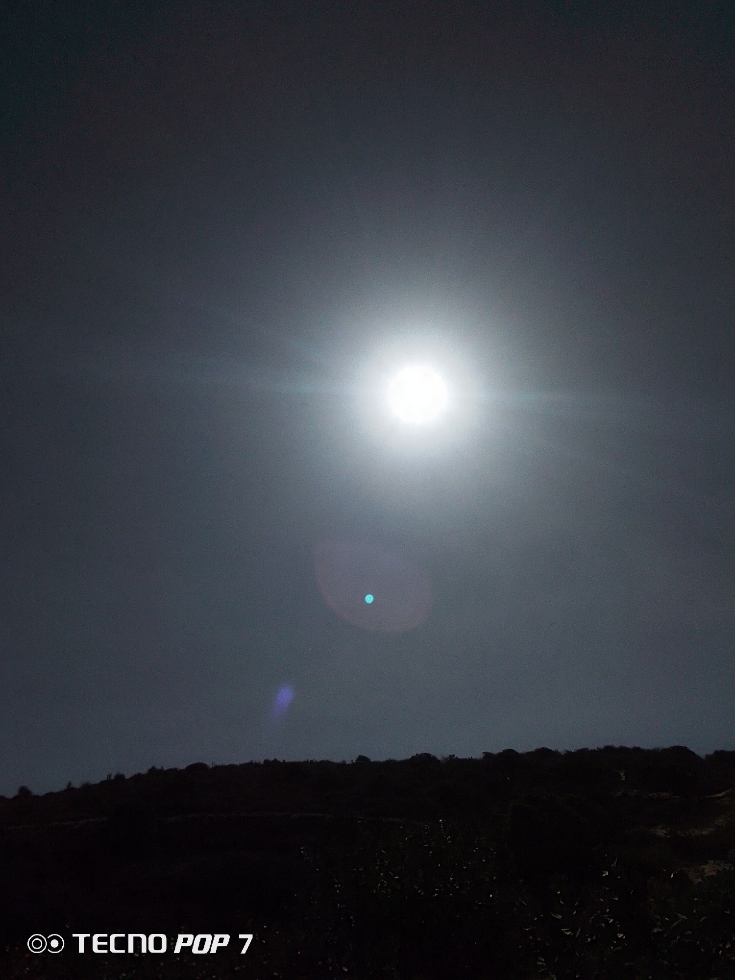 A photograph taken in Pedro González, Venezuela, shows a bright celestial body, possibly the sun, shining against a dark sky. Below, the dark silhouette of a land mass, possibly a mountain or hill, stretches across the lower portion of the image. The words "TECNO POP 7" are visible in the bottom left corner. The light has created lens flare.