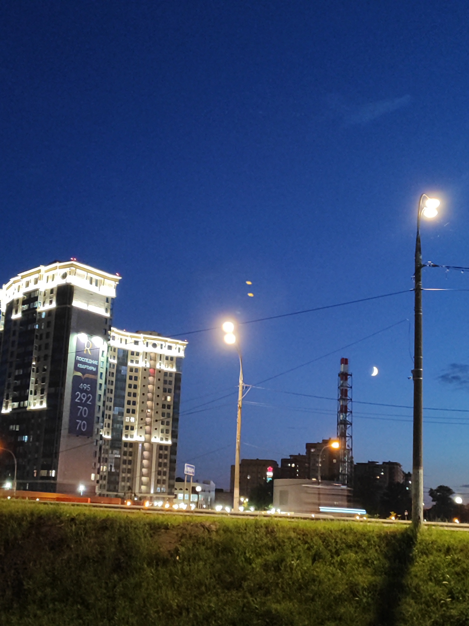 A nighttime exterior shot depicts a city skyline against a dark blue sky. Two modern, illuminated high-rise apartment buildings dominate the left side of the frame, with their windows and decorative rooftop lighting casting a glow. A large banner on the left building displays the text "ПОСЛЕДНИЕ КВАРТИРЫ" and numbers "495 292 70 70 70". In the mid-ground, a tall, red and white striped industrial smokestack stands out, with a crescent moon visible to its left. A street lamp with two lights is positioned in the foreground on the right, illuminating the scene. In the distance, several other buildings and a blurred vehicle with streaking lights suggest ongoing traffic. A grassy slope occupies the bottom portion of the image.