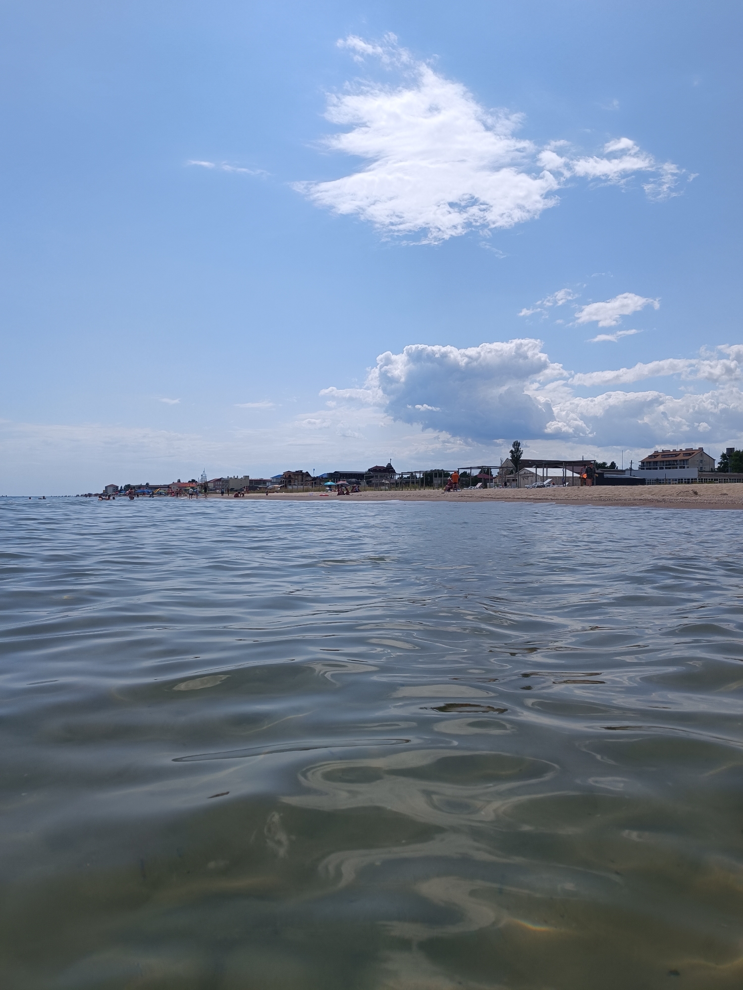 The image depicts a low-angle view of the Black Sea coast in Zatoka, Ukraine. The foreground is dominated by calm, rippling water, with sunlight reflecting off its surface and revealing a murky, shallow seabed. In the midground, a sandy beach is visible, lined with various buildings, including hotels and beach facilities, and dotted with numerous beach umbrellas and sunbeds. Several people are present on the beach, some swimming in the water, others relaxing on the sand. The background features a clear blue sky with scattered white clouds, indicating fair weather. The scene conveys a typical summer day at a popular seaside resort.