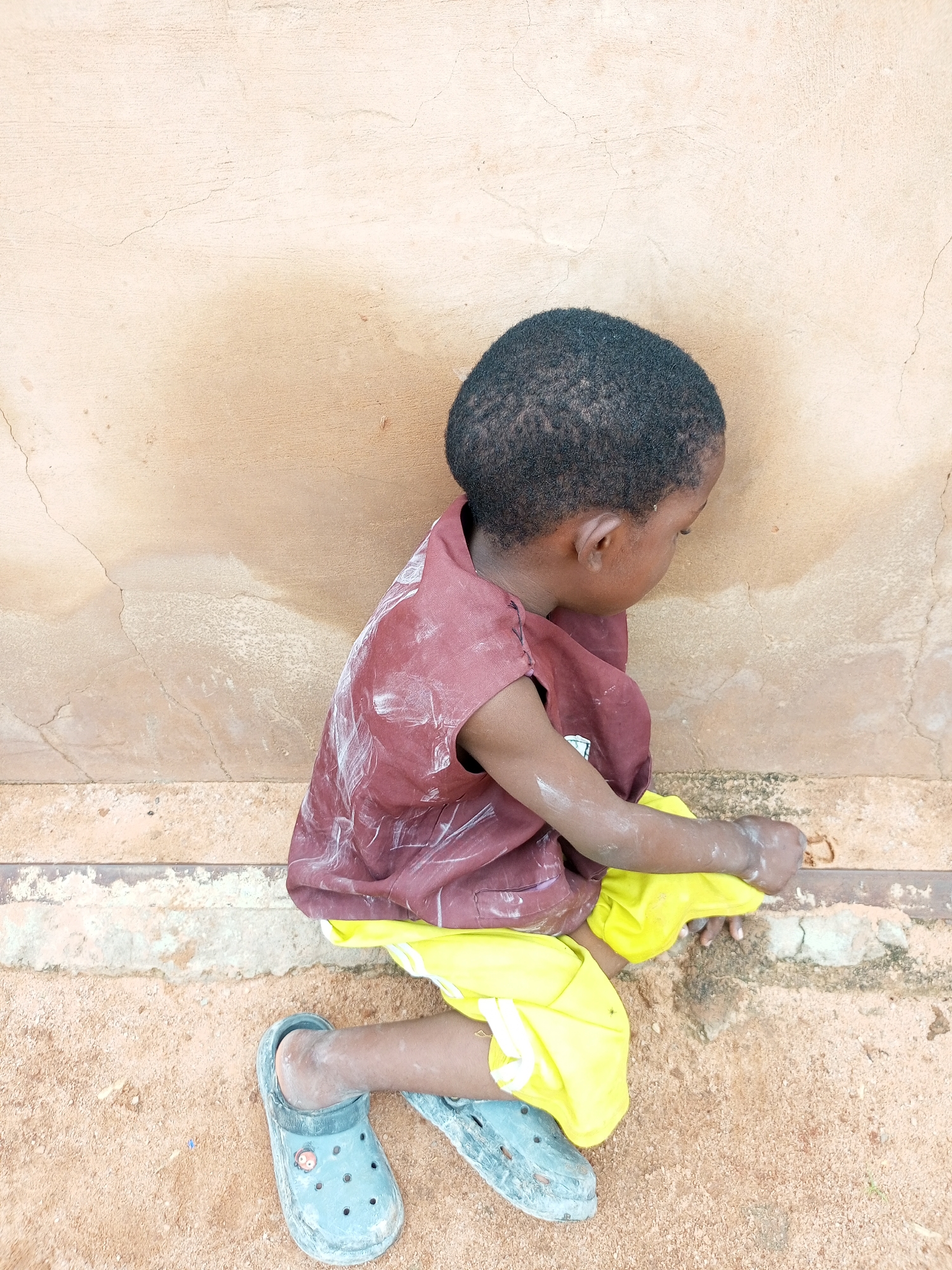 A young child kneels on sandy ground in front of a light brown, textured wall. The child, facing to the right and partially obscured from view, wears a maroon shirt with white smudges and bright yellow shorts. The child's right foot is visible, clad in a gray Croc-style shoe, and the child's hands are engaged with the sand. The environment appears to be outdoors, possibly in an urban setting within Tempure, Nigeria, indicated by the presence of the wall and the ground texture.