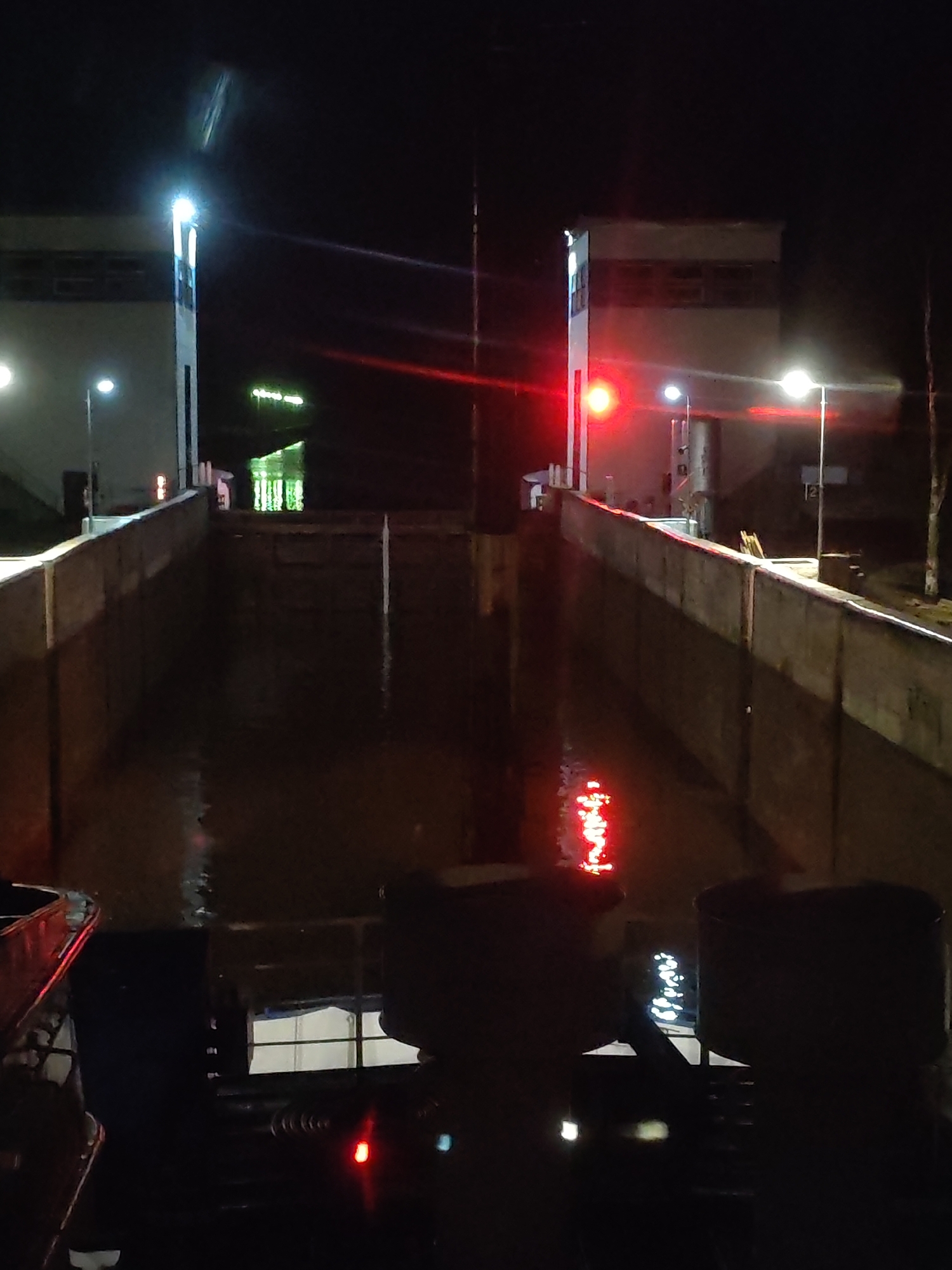 The image shows a night view of a canal lock in an unidentified location. The water is reflecting red and white lights, presumably from structures on either side of the canal. Two control towers stand on opposite sides of the waterway. Each tower has a bright white light at the top and a lower red signal light. The foreground displays the blurred silhouette of the interior of a boat, possibly a tug or pilot boat, with visible equipment. The environment is dark, with ambient lighting on the edges of the waterway and minimal illumination illuminating the canal walls.