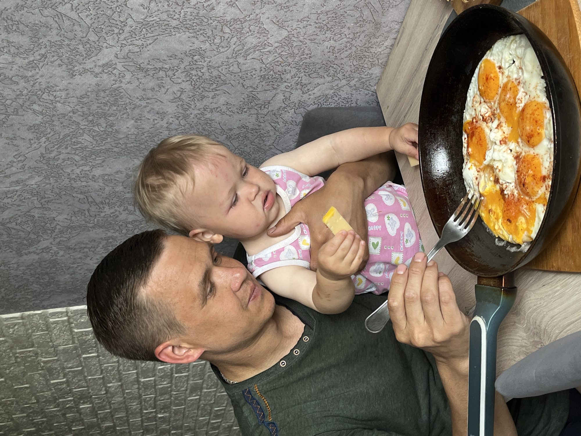 A man holding a toddler is holding a pan with fried eggs. The toddler is holding a small, yellow object. The man is holding a fork and is looking at the pan. The background is a textured wall. The photograph was taken in the city of Chaikovsky, Russia.