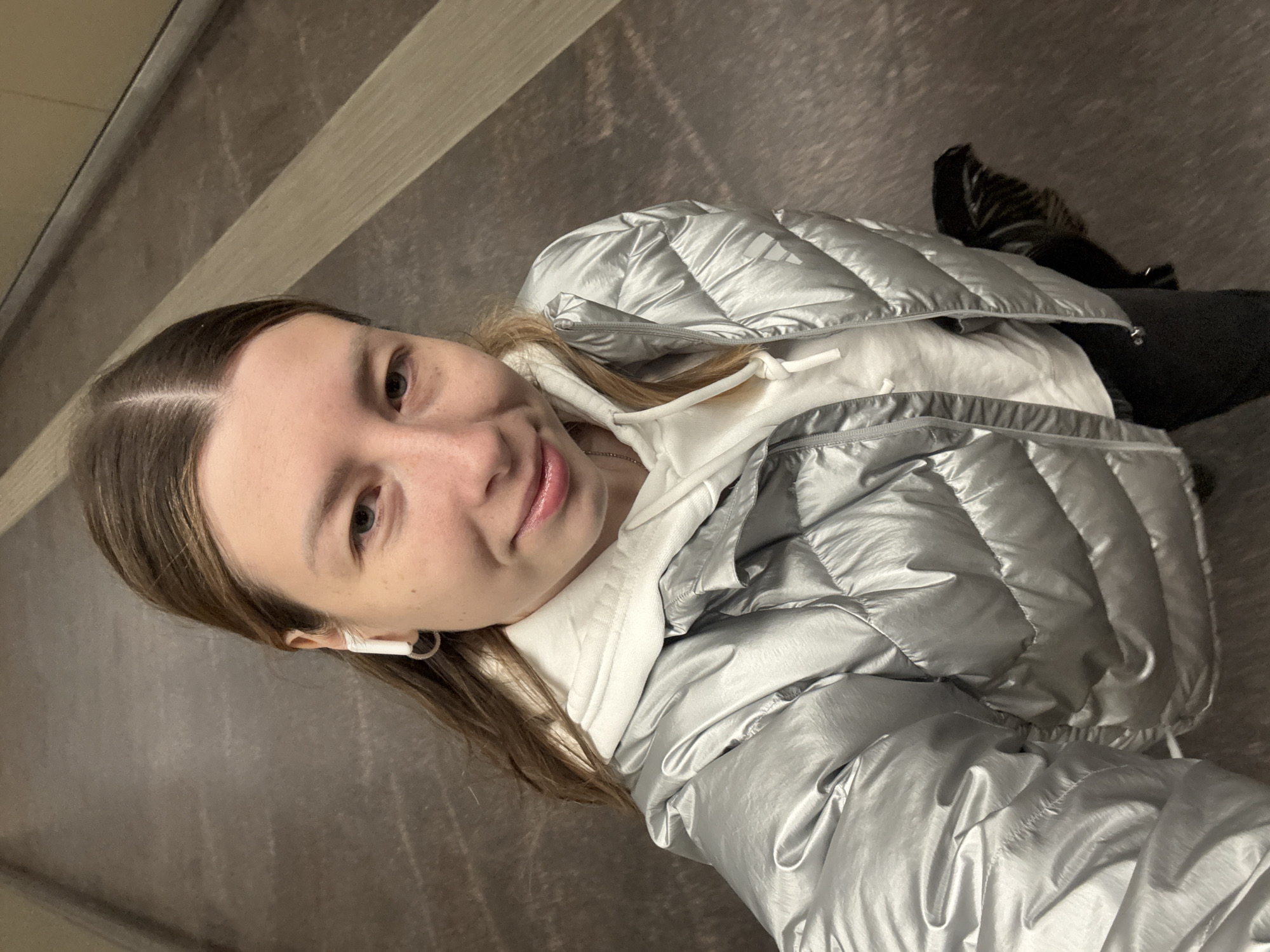 A young woman is captured in a selfie, positioned vertically with her head towards the left side of the frame. She has long, light brown hair parted down the middle, and is looking directly at the camera with a slight smile. Her eyes are open and her expression is pleasant. She wears a white wireless earbud in her left ear.
She is dressed in a silver, metallic puffer jacket over a white hooded sweatshirt. The puffer jacket features distinct quilted sections, and the Adidas logo is subtly visible on her right shoulder in one of the cropped images. A necklace with a small pendant is partially visible beneath her hoodie. The jacket's material appears shiny and reflective.
The background consists of plain, muted gray and brown tones, suggesting an indoor or covered setting, possibly a hallway or public transit area. Linear patterns are visible on the wall or floor behind her, running diagonally. A glimpse of what appears to be a black shoe or bag is visible in the upper right background. The image context indicates the location is Moscow, Russia, though no specific landmarks are visible. The overall mood is casual and relaxed.