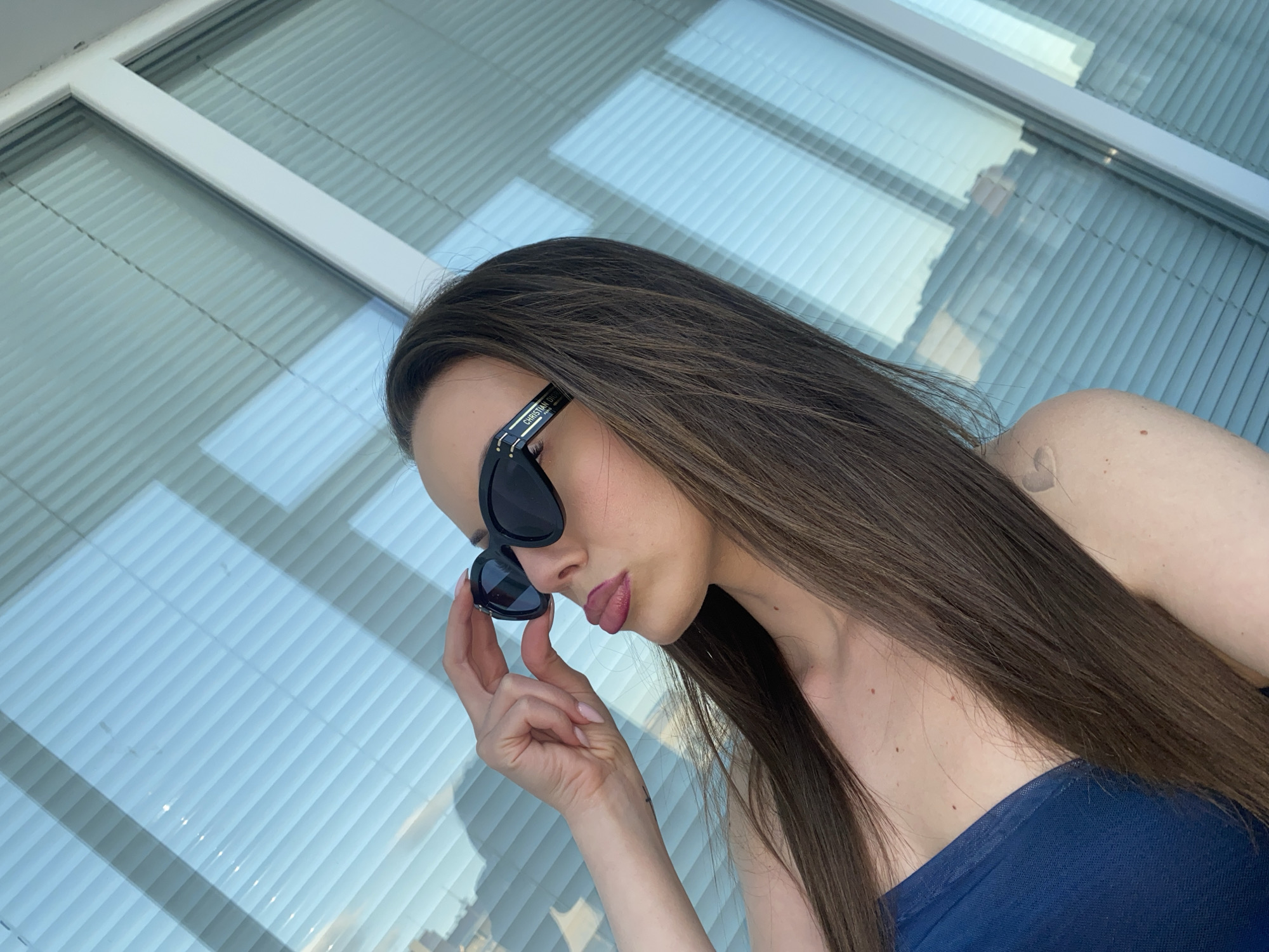 A woman with long, dark brown hair and bold pink lipstick is posing for a selfie against a backdrop of blue blinds. She is wearing black sunglasses with the Christian Dior logo on the arms and a dark blue strapless top. Her left hand is raised, holding the sunglasses up to her face. The blinds are angled, suggesting a window with a view of buildings outside, though the details are obscured by the lighting and the angle of the blinds. The overall scene appears to be indoors, possibly on a balcony or near a window.