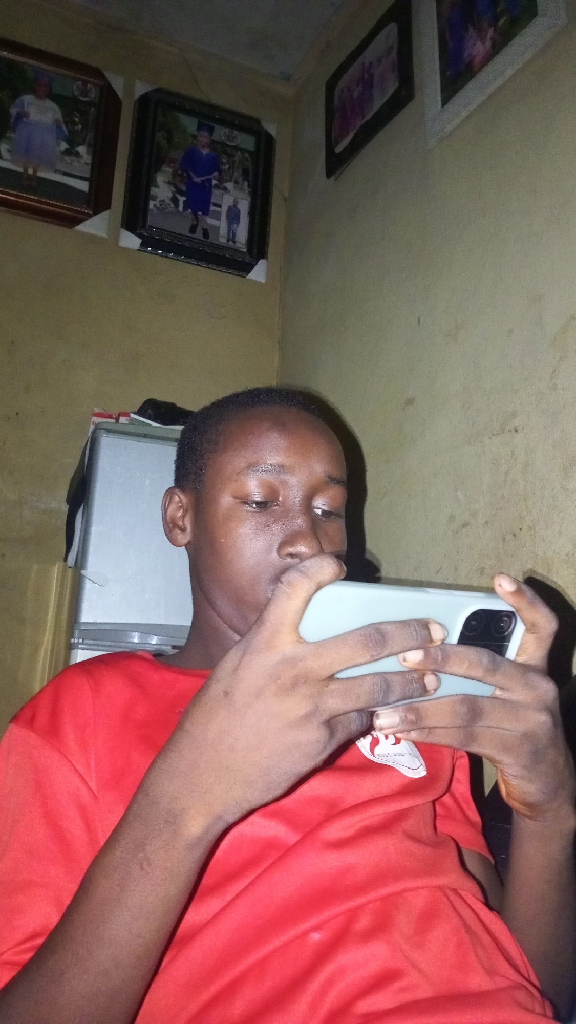 A person with dark skin and short hair is holding a light gray cell phone in both hands. They are looking at the phone's screen. The person is wearing a red t-shirt with a white logo on the front. In the background, there is a white metal cabinet or small refrigerator. On the wall behind the person, there are three framed photographs. The location appears to be a room with yellowish walls. The city and specific location are Unknown, Unknown.