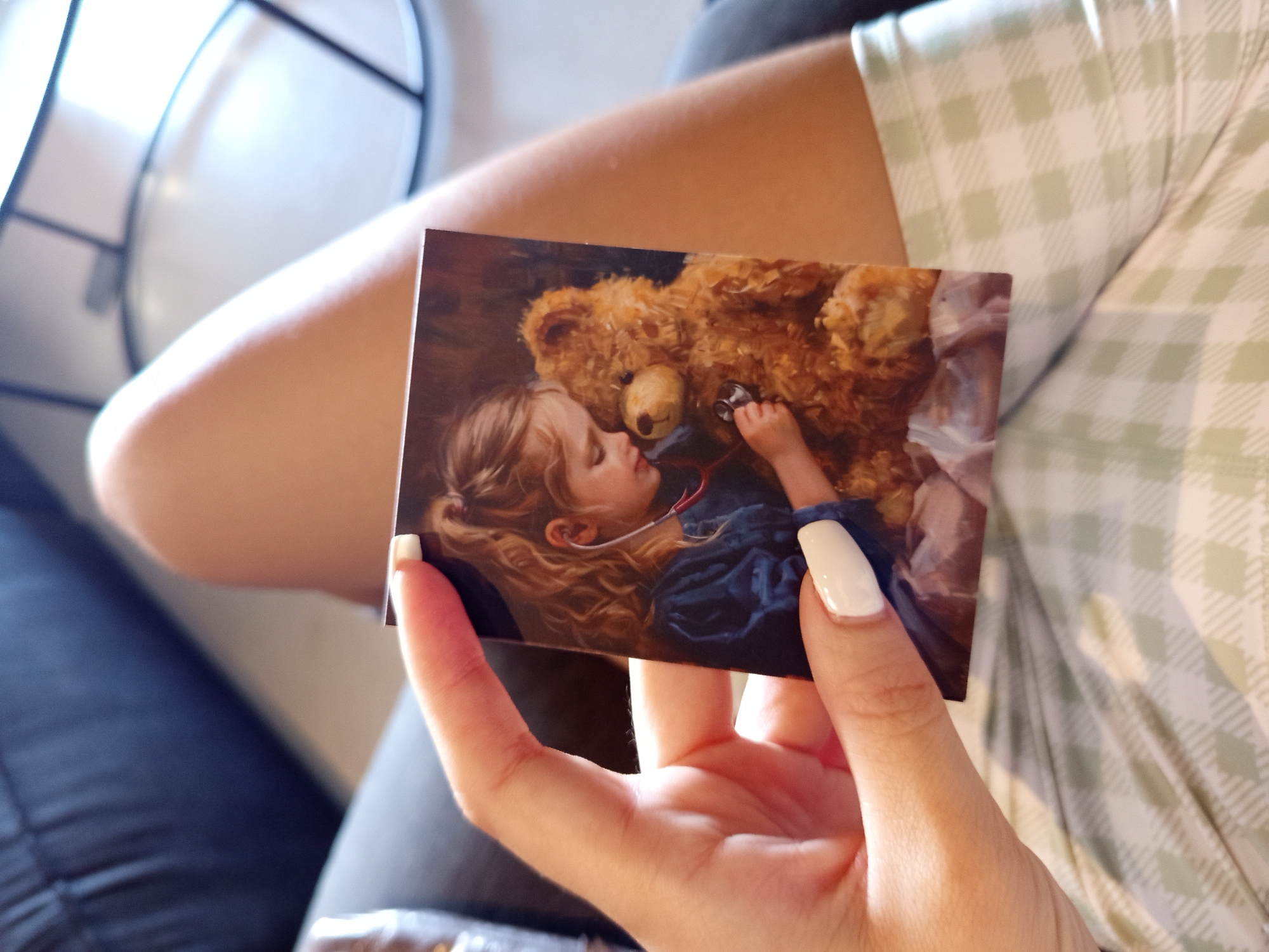 A hand holds a photo of a child examining a teddy bear with a stethoscope. The child has blonde curly hair. The teddy bear is a light brown color. The background of the photo is a dark, reddish-brown. The hand belongs to a person with long, white manicured nails. The person is wearing a light-colored, green-and-white checkered patterned garment. The person is sitting on a dark gray or black upholstered seat. A portion of a black metal chair is visible in the upper-left background. The location is unknown. The location of North Kuta, Indonesia, cannot be verified from this image.