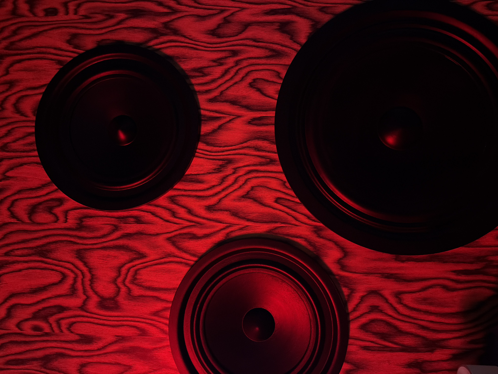 The image shows three black circular speakers mounted on a reddish-brown wood surface. The speakers are arranged as follows: one is in the top left, one is in the top right, and the last one is at the bottom center. The wood surface has a prominent grain pattern. The entire scene is illuminated by a red light. There is no information in the image to suggest its location in the city of Sofia, Bulgaria.