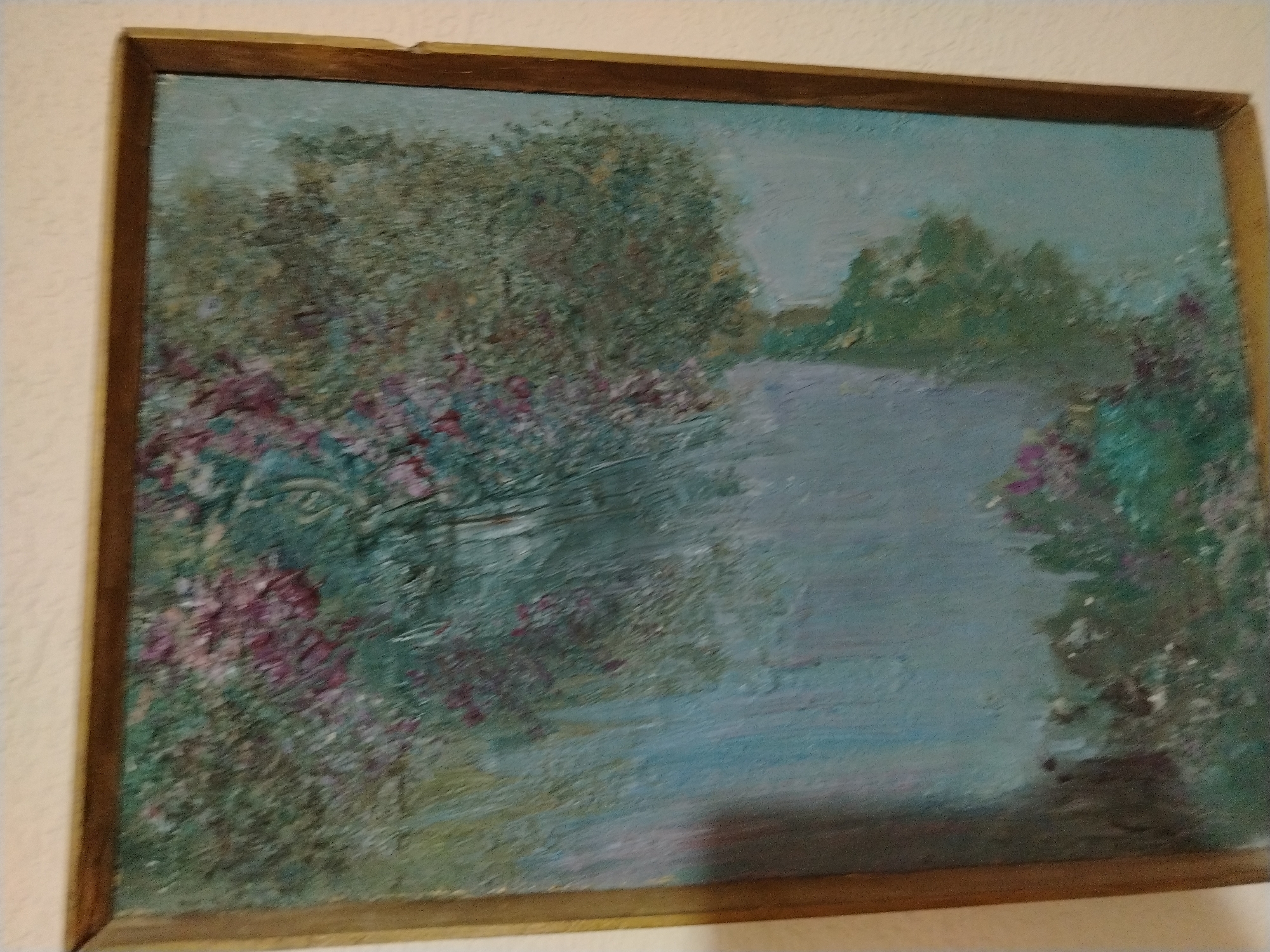 The image shows a painting of a body of water with trees and flowers on the banks. The painting is framed in a simple wooden frame. The style of the painting is impressionistic, with visible brushstrokes and a focus on light and color. The scene appears to be a peaceful landscape, possibly a garden or a park. The specific location in Nukus, Uzbekistan, cannot be determined from the image alone, as it is a generic landscape painting.