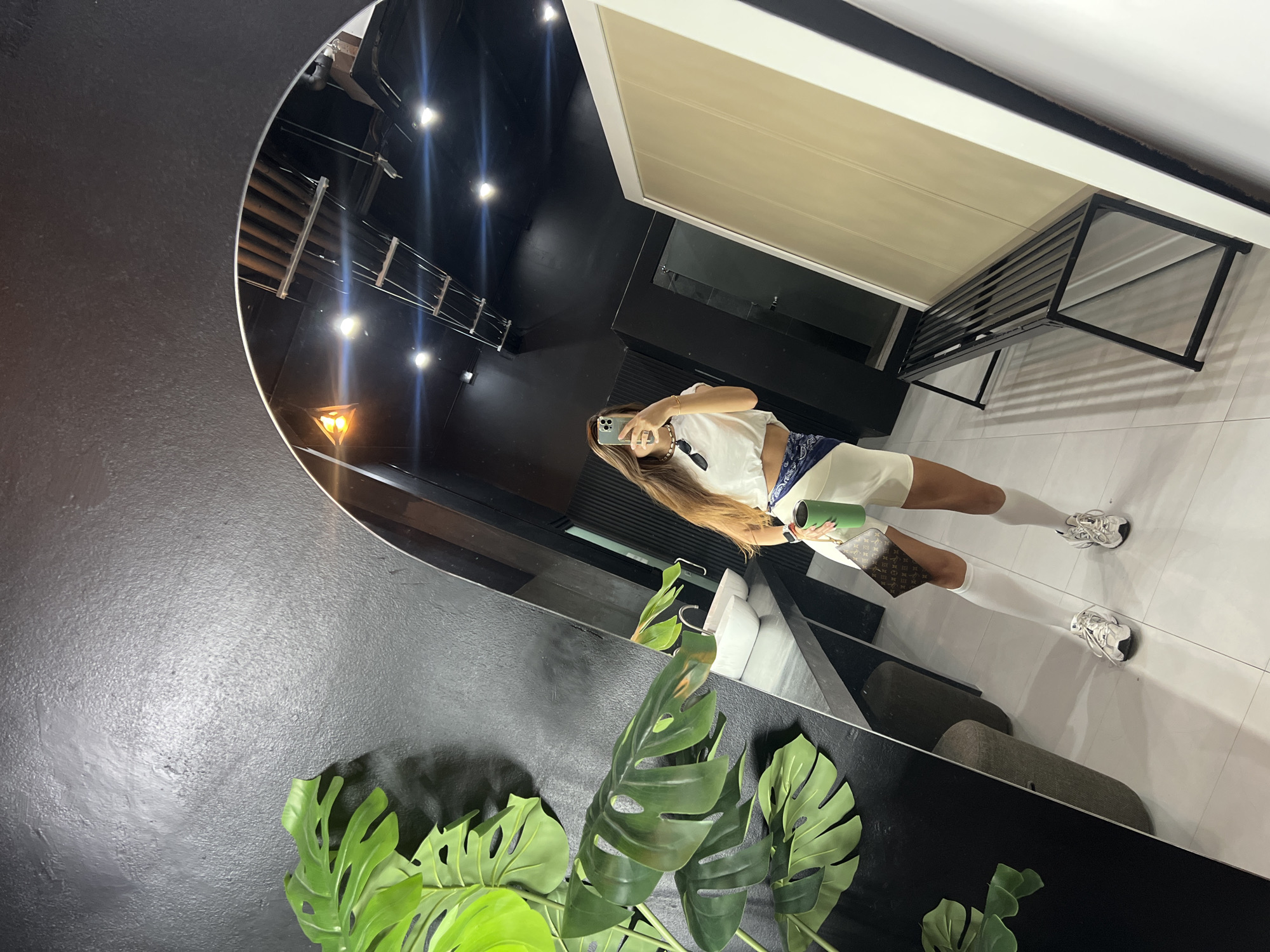 A woman with long, brown hair stands in front of a large, arched mirror, taking a selfie. She is wearing a white crop top, white shorts, white socks, and white athletic shoes. A blue and white bandana is tied around her waist, and she is carrying a green cup and a small brown handbag with a monogram pattern. The mirror reflects a room with black walls and ceiling, a sink, a bench, and a large plant with green leaves. The floor is tiled white. The overall impression is that of a modern, possibly a gym or a locker room.