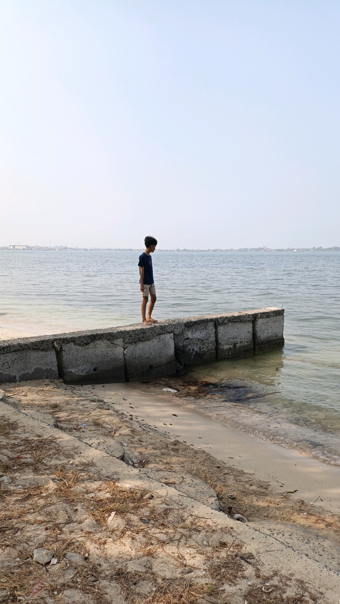 A young boy stands on a concrete seawall at the edge of a sandy beach in Cikal, Indonesia. He is wearing a dark blue t-shirt and light-colored shorts. The boy is facing away from the camera, looking out at the calm, gray water of the sea. The seawall is made of large, rectangular concrete blocks, some of which are broken or weathered. The beach in the foreground is covered with sand, dry grass, and debris. In the distance, a hazy shoreline with buildings and structures can be seen. The sky is clear and pale blue, indicating it may be late morning or early afternoon.