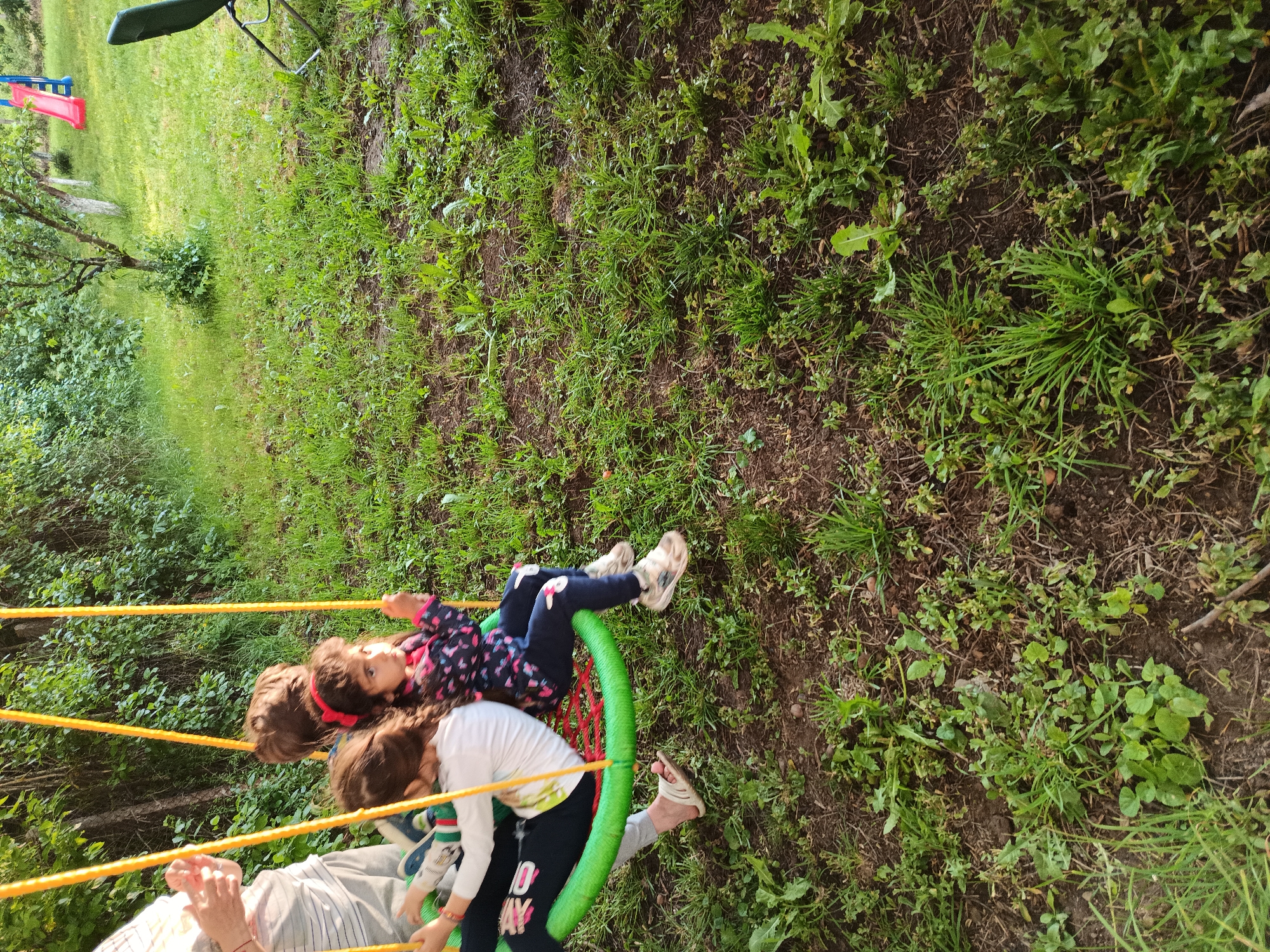 The image depicts an outdoor recreational scene in Kovachevtsi, Bulgaria. Three children and one partially visible adult are present in a garden or backyard setting.
On the left side of the frame, an adult's torso and lower limbs are visible, wearing a light-colored striped or plaid shirt and light-colored pants. Their right hand is positioned near a circular "nest" swing, which features a thick green outer ring and a red netted seating area, suspended by four yellow ropes.
Three children are seated on the swing. The leftmost child, partially obscured, wears a white long-sleeved top and dark pants with white text or patterns; their feet, clad in light-colored flip-flops, are visible. A second child, positioned centrally, is largely obscured, with only the back of their brown-haired head and a portion of their back visible. The third child, on the right, is a young girl with dark, curly hair and a red headband. She is dressed in a dark blue jacket with a colorful pattern, dark blue pants with white appliques on the knees, and light-colored athletic shoes. Her gaze is directed towards the right side of the frame.
The ground is covered with a mixture of green grass, various broadleaf weeds (including dandelions), and patches of exposed dark soil. In the background, a dense treeline composed of green deciduous trees and bushes forms a natural barrier. To the mid-right, a small, sparsely foliated deciduous tree stands on the grassy ground. Further right, a red and blue plastic children's slide is visible, and on the far right edge of the frame, a dark green outdoor chair with metal legs is partially present. The scene is illuminated by natural daylight.