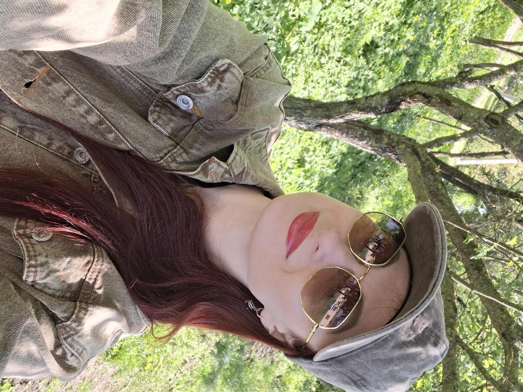 A woman with long auburn hair, wearing a brown denim jacket, a gray cap, and gold-rimmed sunglasses, is lying on her side in a grassy area with trees, possibly in Забалоцкі сельскі Савет, Belarus. She has fair skin and is wearing red lipstick. Her head is tilted towards the viewer, with her face partially obscured by the sunglasses. Her hair is spread out on the jacket, and a small earring with a blue stone is visible in her left ear. The background consists of green grass and tree branches.