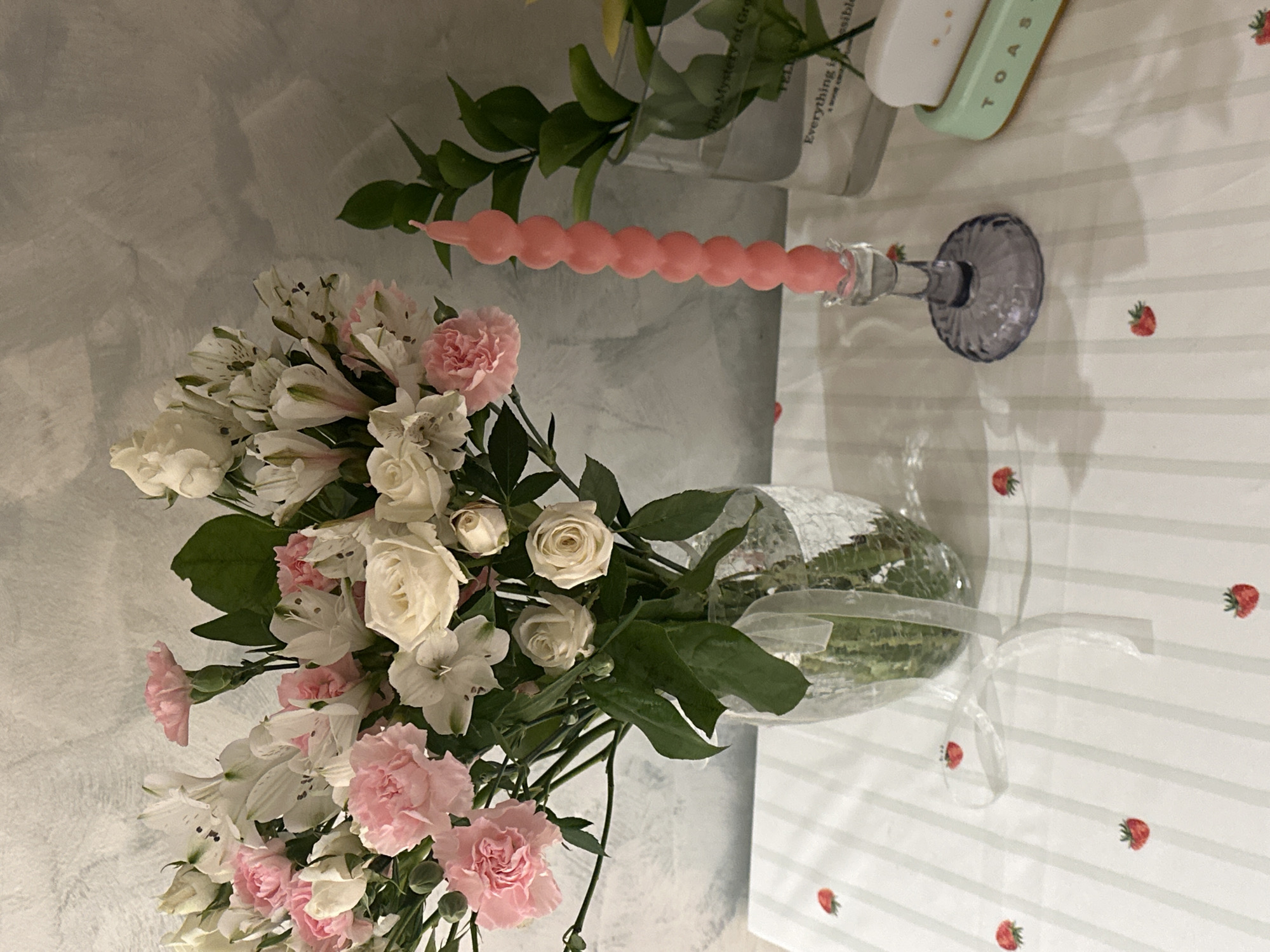 A still life featuring a bouquet of white and pink flowers in a clear vase wrapped in plastic, next to a pink bubble candle in a blue glass candlestick, a clear glass vase with green leaves and yellow flowers, and a white toaster. The scene is set on a white tablecloth with a light gray striped pattern and scattered strawberry motifs. The background includes a light gray wall. The location appears to be indoors. The presence of the toaster and flowers suggests a domestic setting. The city of Šušanj, Montenegro, is not discernable from the image.