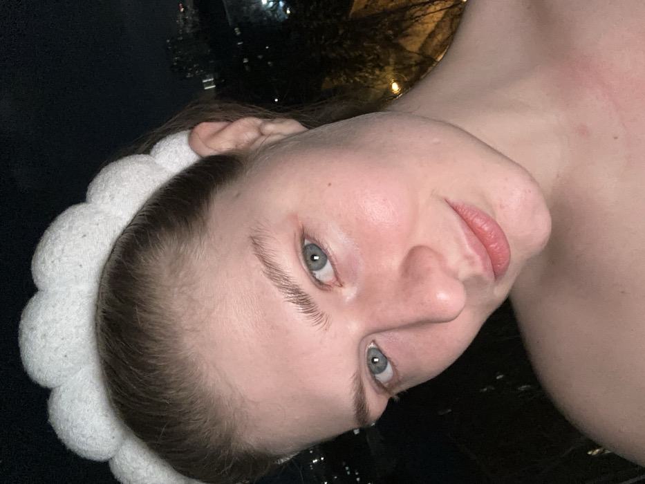 A fair-skinned person with blue eyes, wearing a white, puffy headband, faces the camera. The person has light brown hair visible under the headband. The background appears dark, possibly nighttime, with blurred lights suggesting a city setting. The person's skin appears smooth and well-lit, with a hint of blush on the cheeks. The image seems to be taken from a slightly elevated angle, capturing the person's face and upper body.