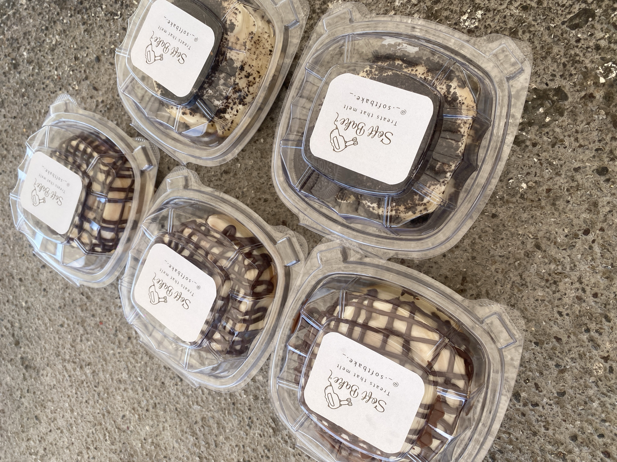 The image shows five transparent plastic containers of desserts, arranged on a textured gray surface, likely concrete or stone. Each container has a white, square label affixed to the top, which reads "Soft Bake Treats that melt @_.softbake._" in a stylized font, accompanied by a cartoon illustration of a hand holding a mixing utensil. The desserts inside appear to be different variations of a creamy filling, with some drizzled with chocolate and others containing cookie pieces. The background is plain and indistinct, suggesting the scene is focused on the desserts themselves.