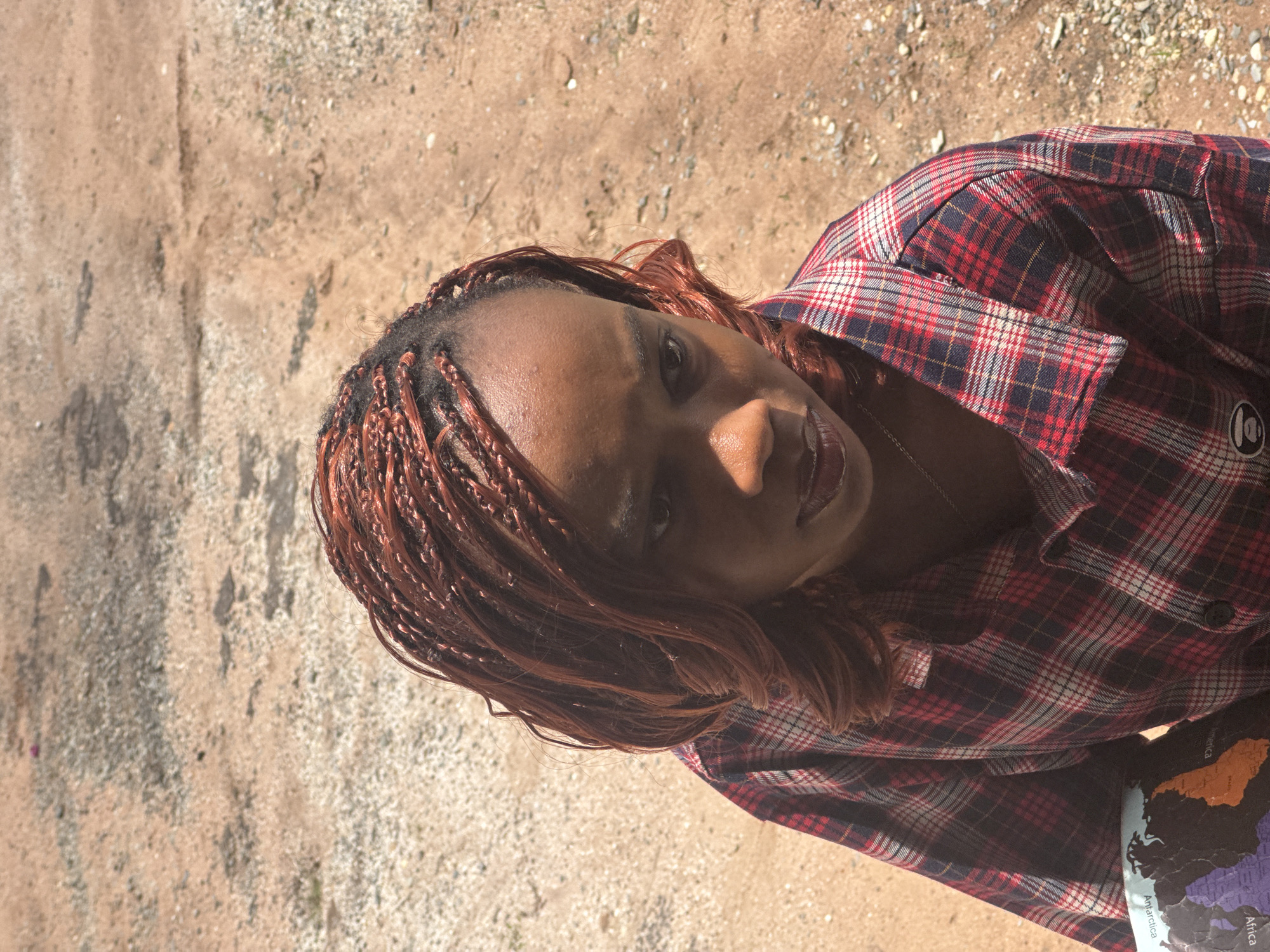 An overhead shot shows a young Black woman with braided reddish-brown hair looking down. She is wearing a red, blue, and white plaid shirt and a necklace. She is holding a globe with a map on it in her left arm. The background is a dirt and gravel surface.