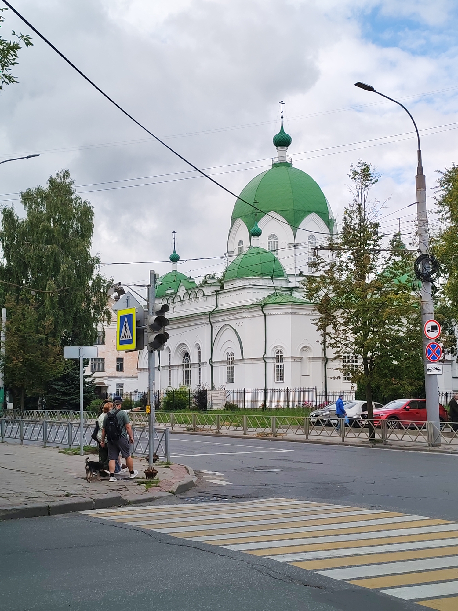 The image depicts an outdoor scene in Bol'shie Mkhi, Russia. A white building with green domes, likely a church, is the focal point. Several trees are visible, with lush green foliage. A road with a pedestrian crossing is in the foreground, with a sidewalk adjacent to it. On the sidewalk, two people and a small dog are present. One person is pointing off-camera. Traffic lights and street signs are also visible. Several cars are parked or driving on the road. The sky is overcast with patches of blue.