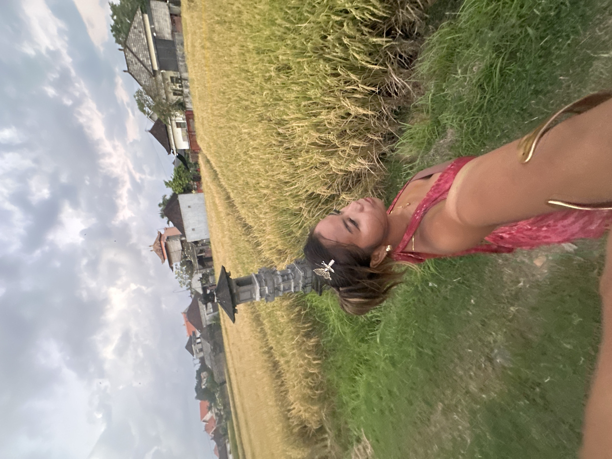 The image shows a person lying in a field of tall grass, possibly rice paddy, in Sukawati, Indonesia. The person is wearing a red dress, jewelry and a hair clip. In the background, there are buildings and houses typical of the region, including what appears to be a traditional Balinese structure. The sky is overcast. The field transitions from tall, golden grass to shorter, green grass near where the person is lying.