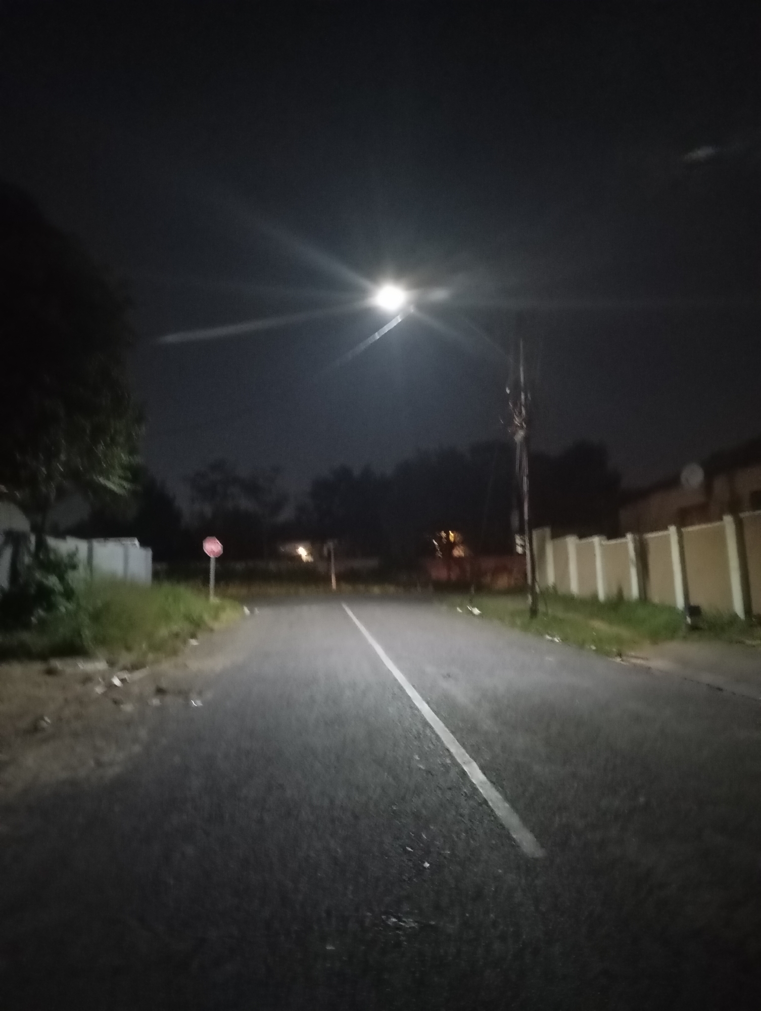 A night scene depicts a road in Johannesburg, South Africa. The road is asphalt, with a white line marking the center. A streetlight illuminates the road, creating a bright spot with light rays. The road is flanked by grassy areas and fences. A stop sign is visible on the left side of the road. An electric pole is on the right side. The sky is dark and cloudy.
