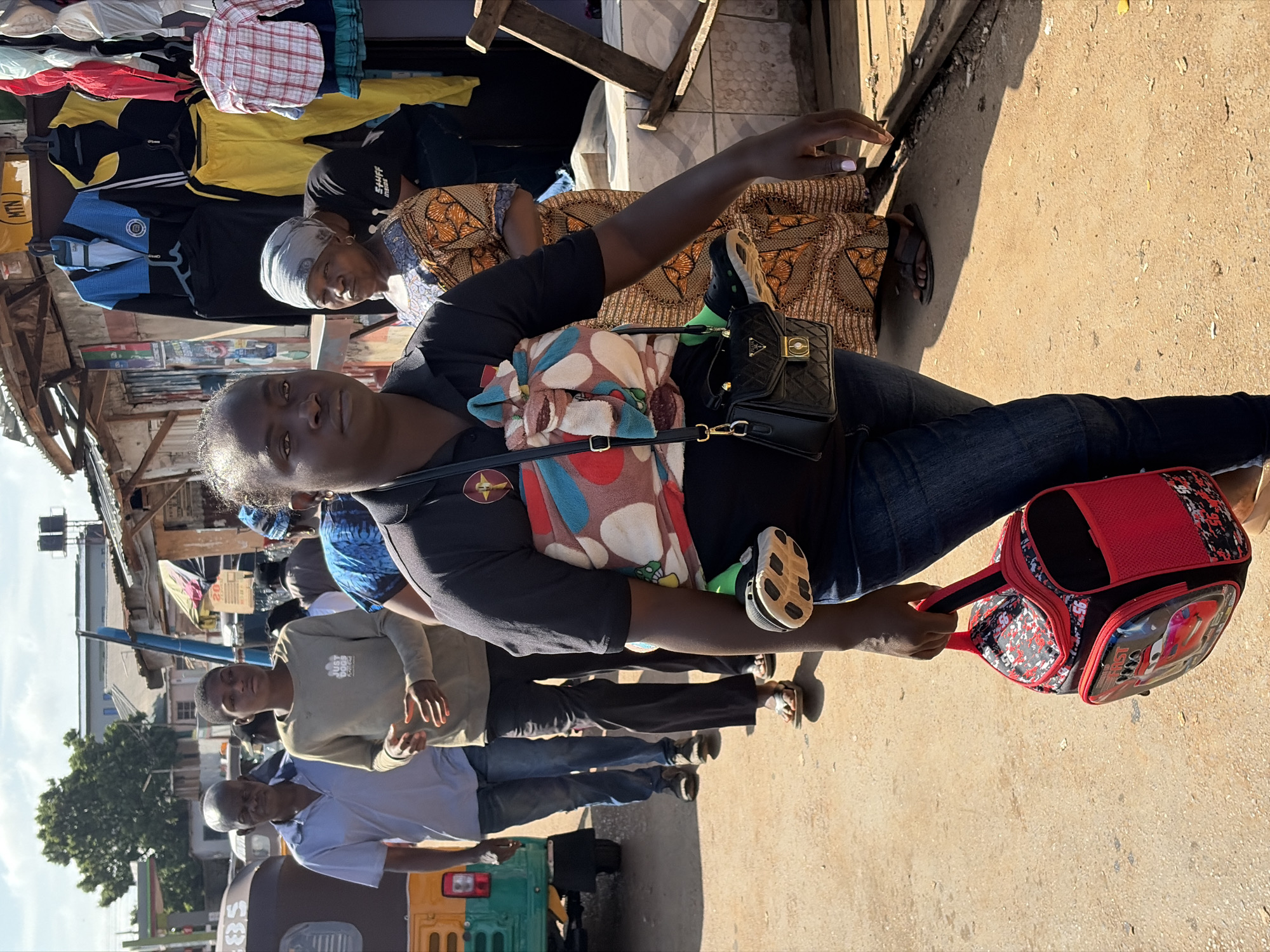 In Gura Top, Nigeria, on a sunlit street, a woman is walking while carrying a baby wrapped in a colorful cloth carrier. She has a black quilted handbag with a gold clasp slung over her shoulder and is holding a red and black backpack with a "Cars" theme in her right hand. She is wearing a dark t-shirt and blue jeans. In the background, other individuals are walking, and a three-wheeled vehicle is visible. Shops line the street, displaying various clothing items on racks, including tracksuits and shirts. The ground is dusty and appears to be a dirt road.