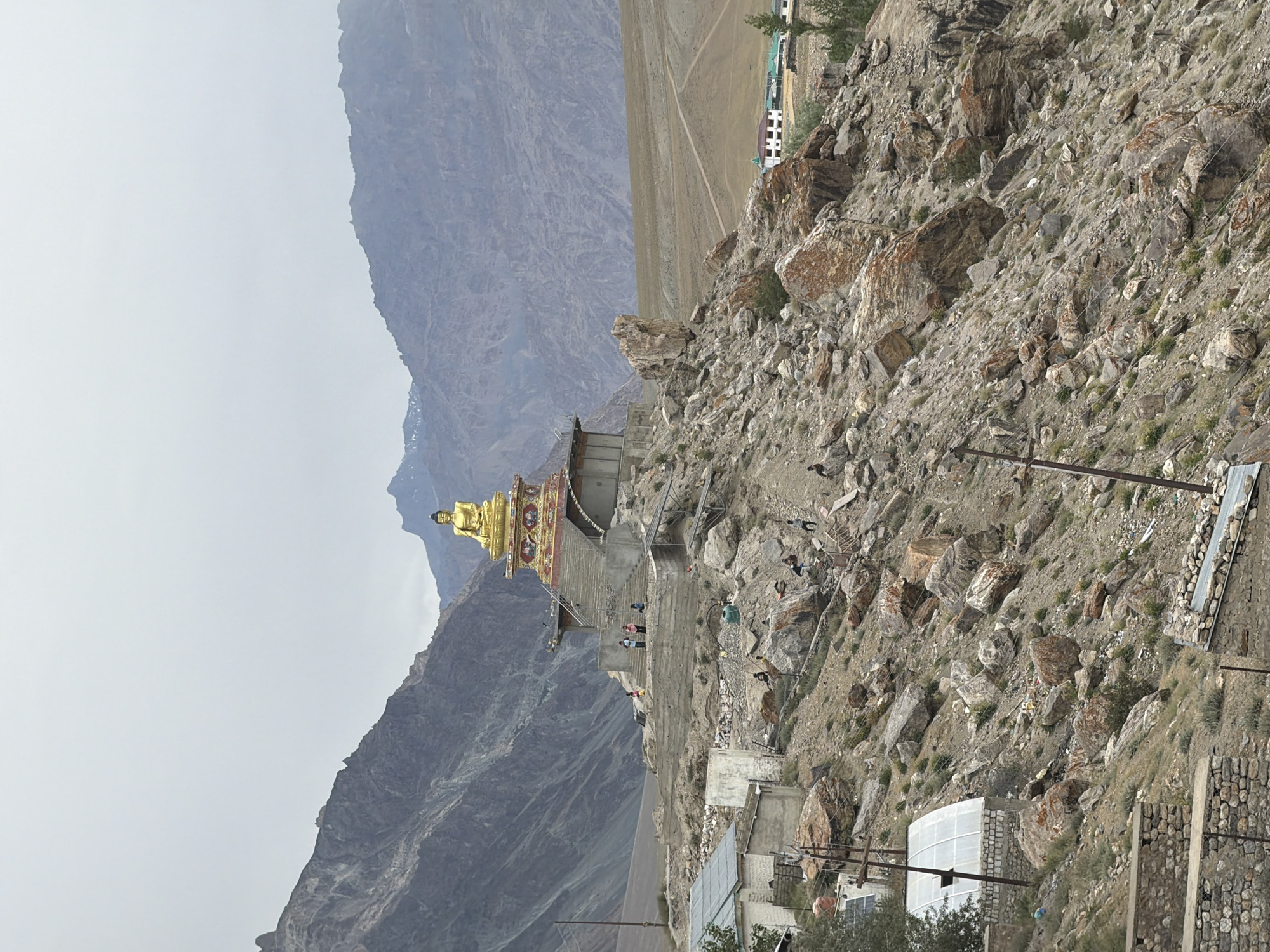 This is an outdoor, vertical, eye-level shot of the ancient Diskit Monastery in Padum, India, on an overcast day. The main subject is a large, golden Buddha statue situated on a raised platform. The statue is seated and appears to be made of polished metal. It is supported by a tiered, red and gold structure with ornate carvings. A wide staircase leads up to the platform.
The monastery is perched on a rocky, arid hillside with sparse vegetation. The terrain is characterized by large boulders and gravel. In the foreground, there are several stone buildings with corrugated metal roofs, and what appears to be a greenhouse-like structure. Some people, dressed in casual clothing, are visible on the steps of the monastery and scattered on the hillside.
In the background, rugged, barren mountains dominate the landscape, with the sky a uniform pale gray. Further in the distance, across a flat, tan-colored plain, a cluster of white buildings with green roofs is visible, possibly part of the village or another monastery. The overall impression is one of a remote, mountainous religious site.