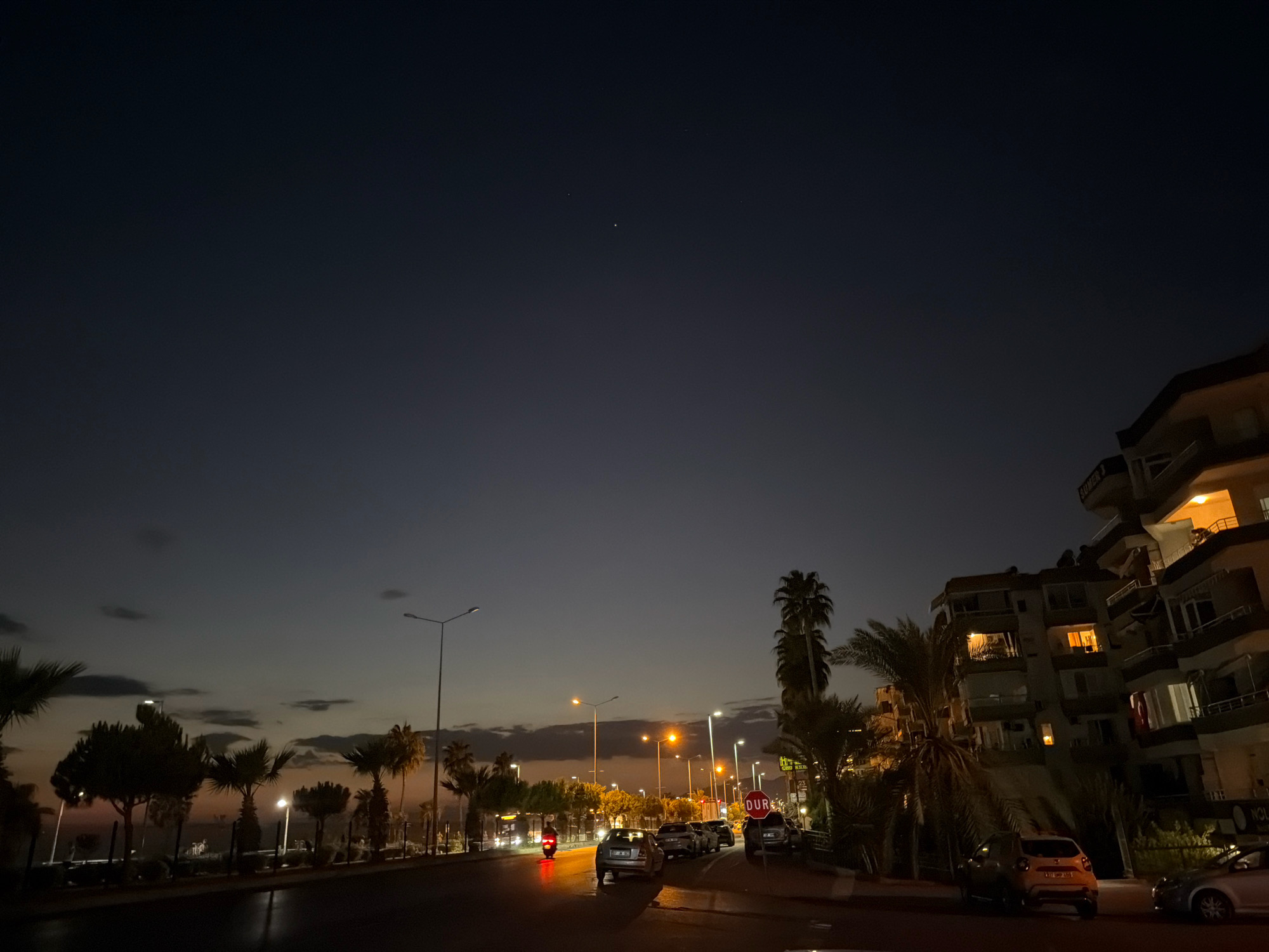 The image depicts a street scene in Alanya, Turkey, at dusk. The sky is a deep blue, transitioning to a faint orange glow near the horizon. Several streetlights are illuminated, casting a warm light on the wet asphalt road, suggesting recent rain. Palm trees line the street, some with their fronds silhouetted against the sky.
On the right side of the road, there are multi-story apartment buildings with illuminated windows, indicating residents are inside. Cars are parked along the street and some are in motion. One car in the foreground is a light-colored hatchback, and behind it, a dark sedan is visible. Further down the road, a motorcycle with its brake lights on is moving away from the camera. A red "DUR" (Stop) sign is prominently displayed on the right side of the road, near the buildings.
In the distance, more streetlights and building lights are visible, creating a sense of urban activity. The overall atmosphere is tranquil yet lively, characteristic of a city evening.