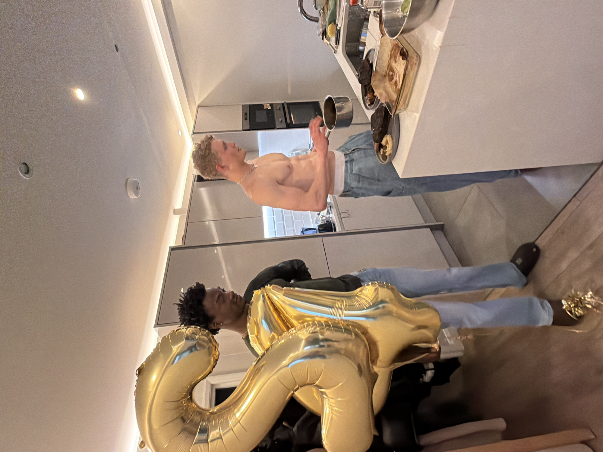 The image depicts two men in a modern, well-lit kitchen and dining area in London, United Kingdom, likely celebrating a 25th birthday.
On the left side of the frame, a man with fair skin and light curly hair stands shirtless, wearing light blue jeans and a white underwear band. He is positioned in the kitchen section, holding a small silver pot or pan with both hands, appearing to be cooking or preparing food. A silver chain is visible around his neck. The kitchen features sleek, light-colored cabinets and integrated appliances, including an oven and microwave, with a clean, contemporary design.
To the right, a second man with darker skin and dark curly hair is seen holding large, gold foil balloons shaped into the numbers "2" and "5," indicating a celebration for a 25th birthday. He is dressed in a dark long-sleeved top and light blue jeans, matching the attire of the first man. His expression suggests he is smiling or laughing, contributing to a jovial atmosphere.
The kitchen counter displays several food items, including a baking dish with what appears to be roasted meat and another plate with dark food and a lighter side dish. A kitchen sink with a modern faucet is also visible. The ceiling features recessed lighting, and strip lighting illuminates the area under the cabinets. A smoke detector is mounted on the ceiling. The overall lighting suggests an indoor evening setting for a casual, celebratory event.