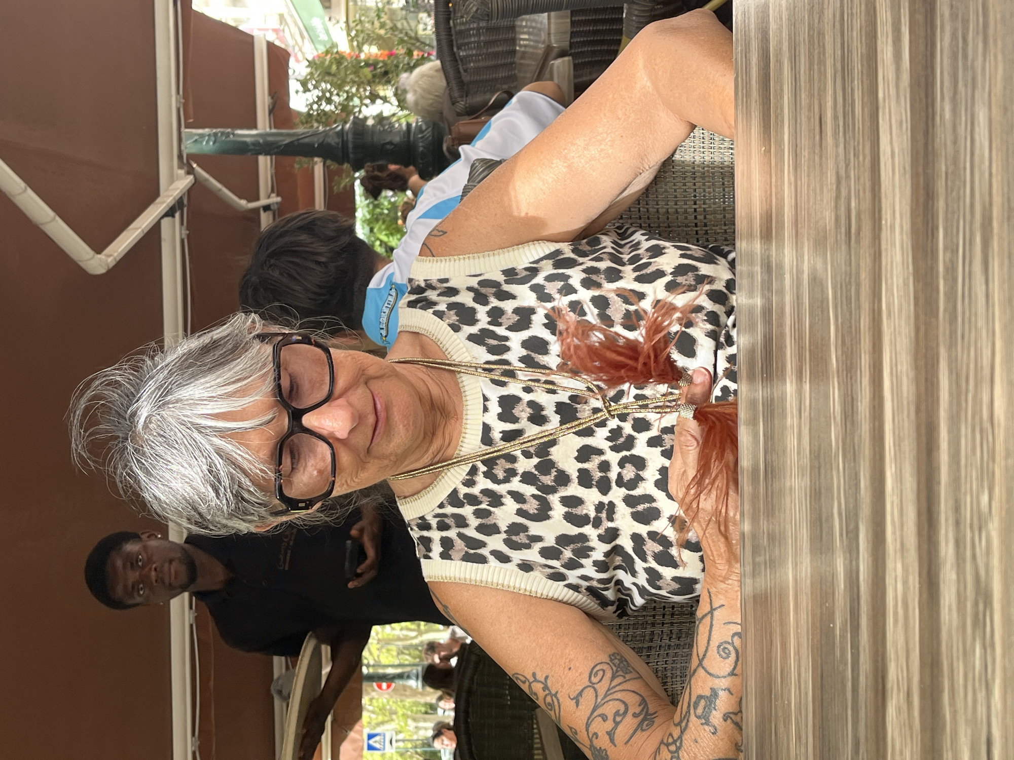 An elderly woman with grey hair pulled up in a bun, wearing large black-rimmed glasses and a leopard print sleeveless top, is seated at a wooden table. Her left arm, adorned with tattoos, rests on the table. She holds a brown feathered object in her hands. In the background, a Black waiter in a black shirt holds a tray and a phone, standing under a brown awning. Another person in an Argentinian soccer jersey is seated behind the woman. The scene appears to be an outdoor cafe in Montélimar, France, indicated by the presence of street signs and buildings visible in the distance through the background foliage.