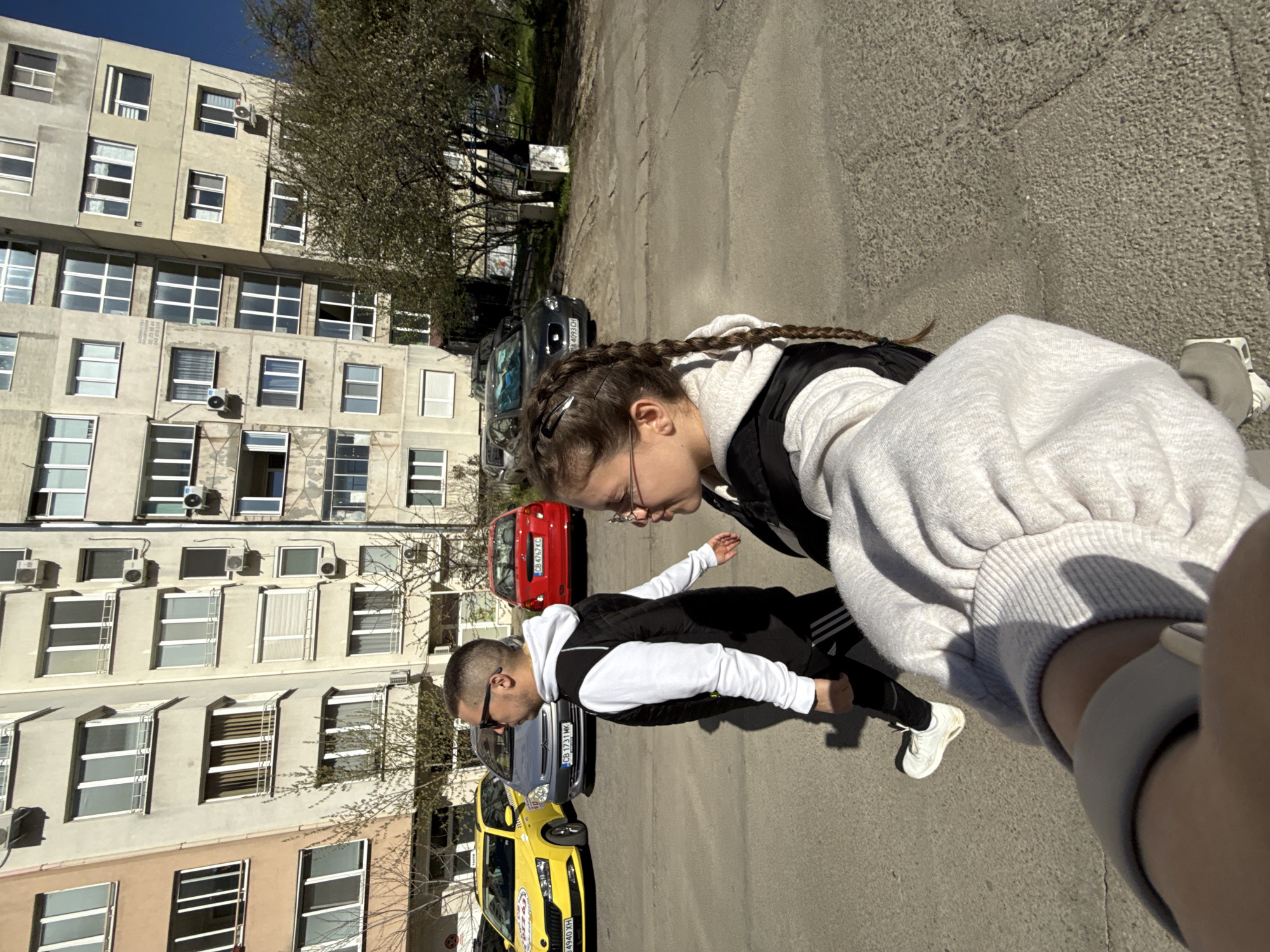 This is a low-angle, outdoor photograph taken on a sunny day in Sofia, Bulgaria. The image captures two people, likely a selfie taken by one of them. In the foreground, a young woman with braids and glasses is visible. She is wearing a light gray hooded sweatshirt and a dark vest. A fitness tracker is visible on her wrist. A man is standing slightly behind her. He is wearing a white hooded sweatshirt, black vest, black pants with Adidas stripes, and sunglasses. They appear to be walking or standing on a street in a residential area.
Several parked cars are visible behind them, including a yellow taxi with the license plate CB 4940XH, a gray car with the license plate CB 1731MK, and a red car with the license plate CB 4747KC. A tall, beige apartment building with numerous windows and air conditioning units is in the background. Bare tree branches are also visible against the building. The street is made of asphalt, and the lighting indicates that the photograph was taken during the day.