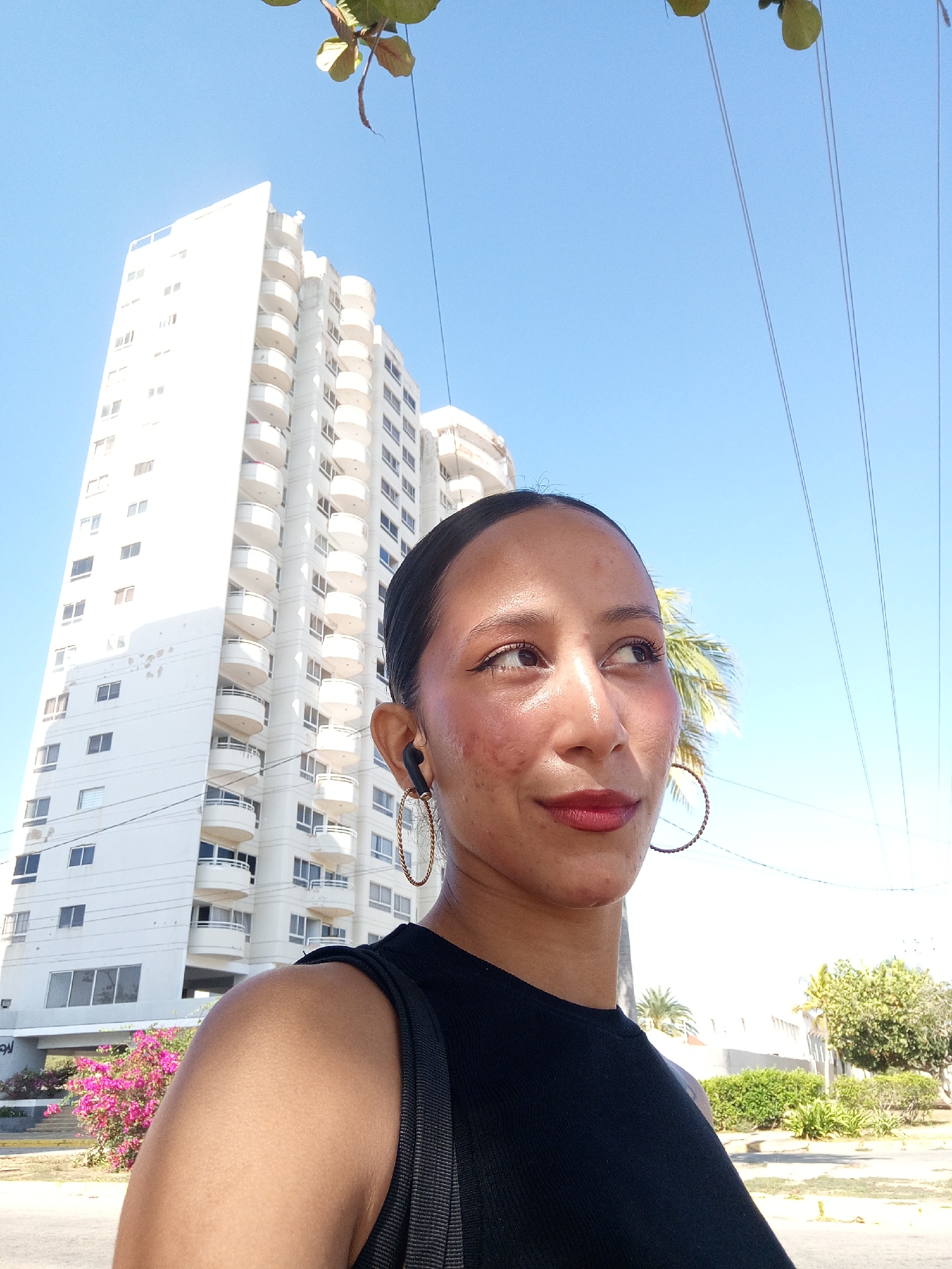 A young woman stands outside in Porlamar, Venezuela. She is wearing a black top and gold hoop earrings. She has dark hair pulled back and appears to be wearing earbuds. A tall white building is behind her to the left, and various trees and plants are in the background to the right. The sky is bright blue.