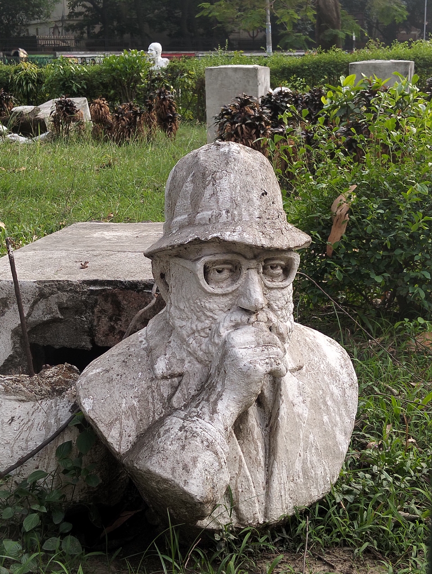 A close-up shot shows a weathered concrete bust of a man with glasses and a hat, his hand resting on his chin in a pensive pose. The bust sits atop a broken concrete base amid overgrown grass and foliage. In the background, there are more concrete structures and greenery, suggesting a park or garden setting. The overall appearance is one of neglect and disrepair.
Based on visual elements, this image likely depicts a sculpture or monument within the Dhaka University campus, specifically the "Sculpture of Intellectuals" located near the TSC (Teacher-Student Centre).