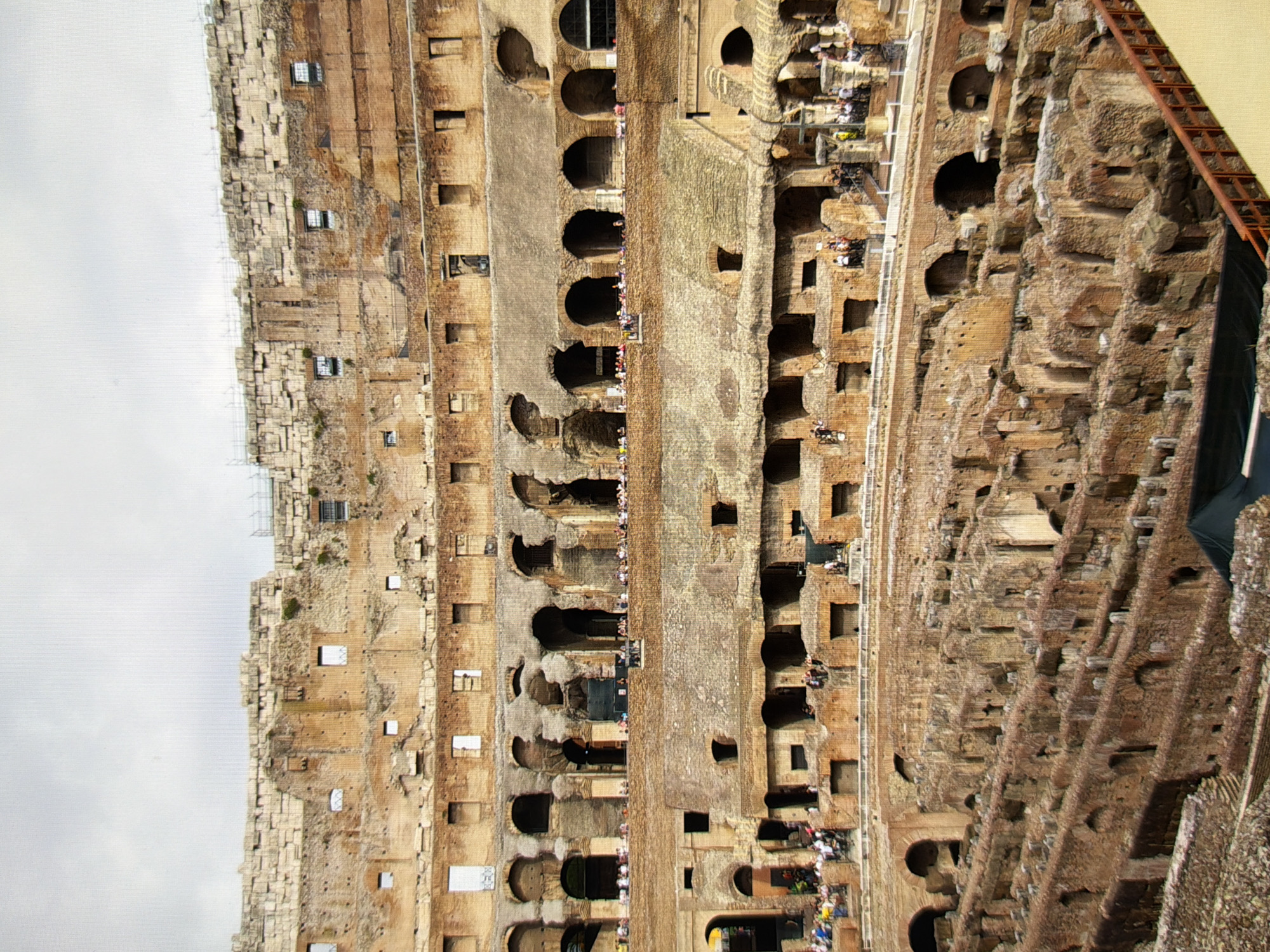 The image shows a view of the Colosseum in Rome, Italy. The ancient Roman amphitheater is made of stone and brick, with multiple levels of arches and seating. Tourists are visible on the lower levels, walking and observing the structure. The sky is overcast. The image does not depict Kyiv, Ukraine.
