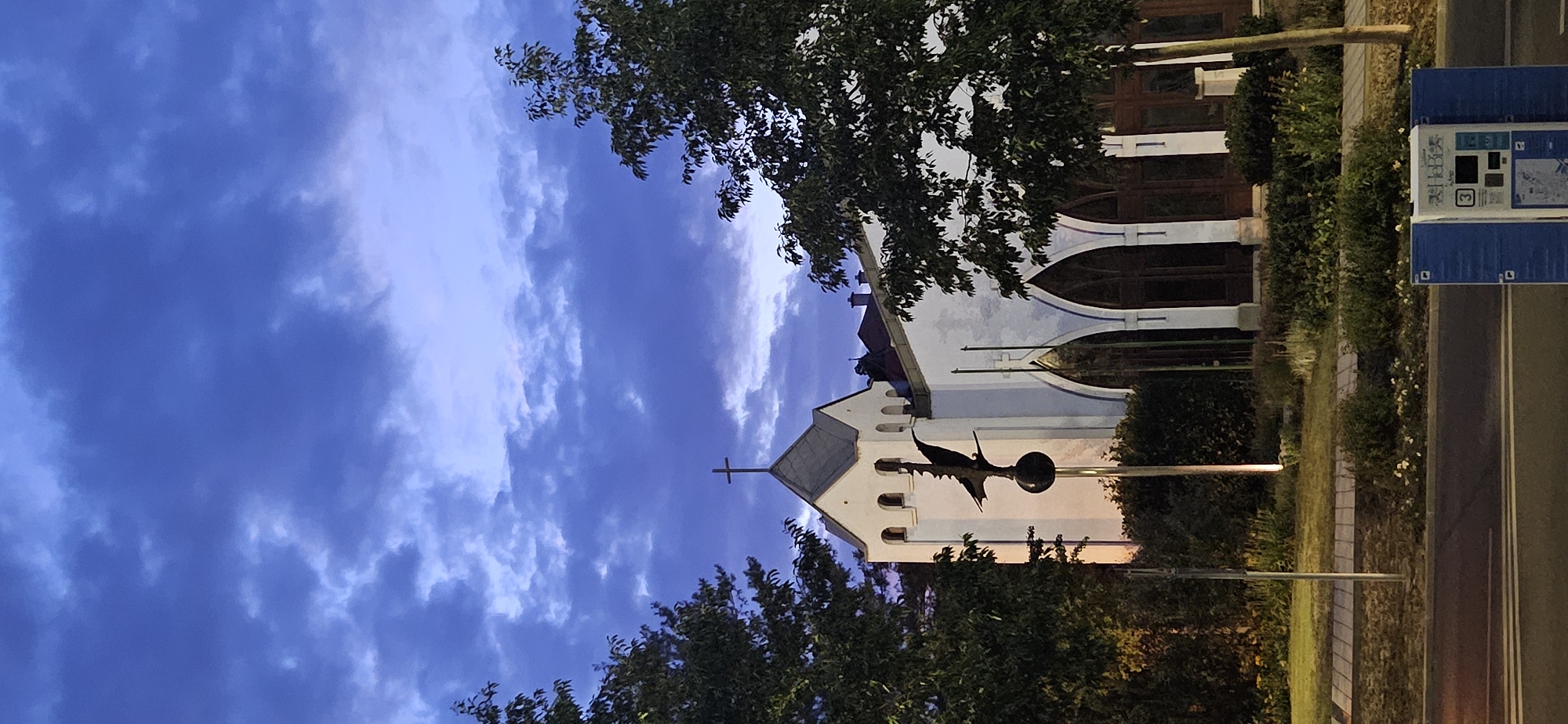An exterior, medium shot captures a church in Hévíz, Hungary, under a twilight sky filled with dramatic, dark blue clouds. The church is white with a grey, gabled roof and a cross at its apex. A sculpture of a bird on a sphere atop a pole stands in front of the church. The church has arched windows and doors. Trees flank the building, and a patch of grass with a sidewalk separates it from a road in the foreground. A blue ticket machine is visible on the sidewalk. The scene is lit by artificial lights, casting a warm glow.