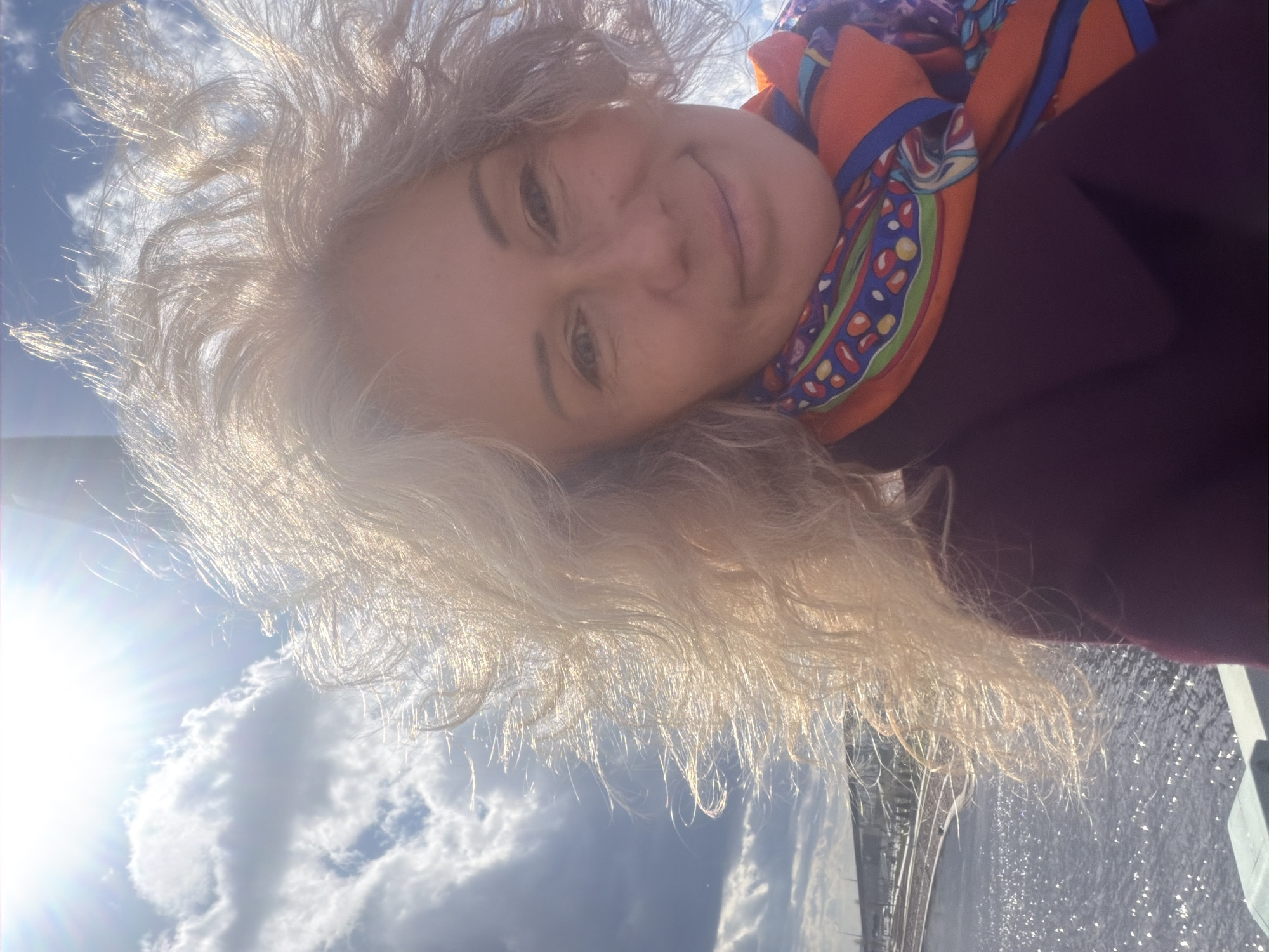 A selfie-style photograph shows a woman with fair skin and graying, curly hair. She wears a colorful scarf with orange, blue, green, and red patterns. She also wears a dark purple coat. The background reveals a sunny sky with white clouds and a body of water, likely a canal or river, with the sun reflecting on the water's surface. A bridge is visible in the distance. Based on the architectural features and waterways, the location is likely Saint Petersburg, Russia.