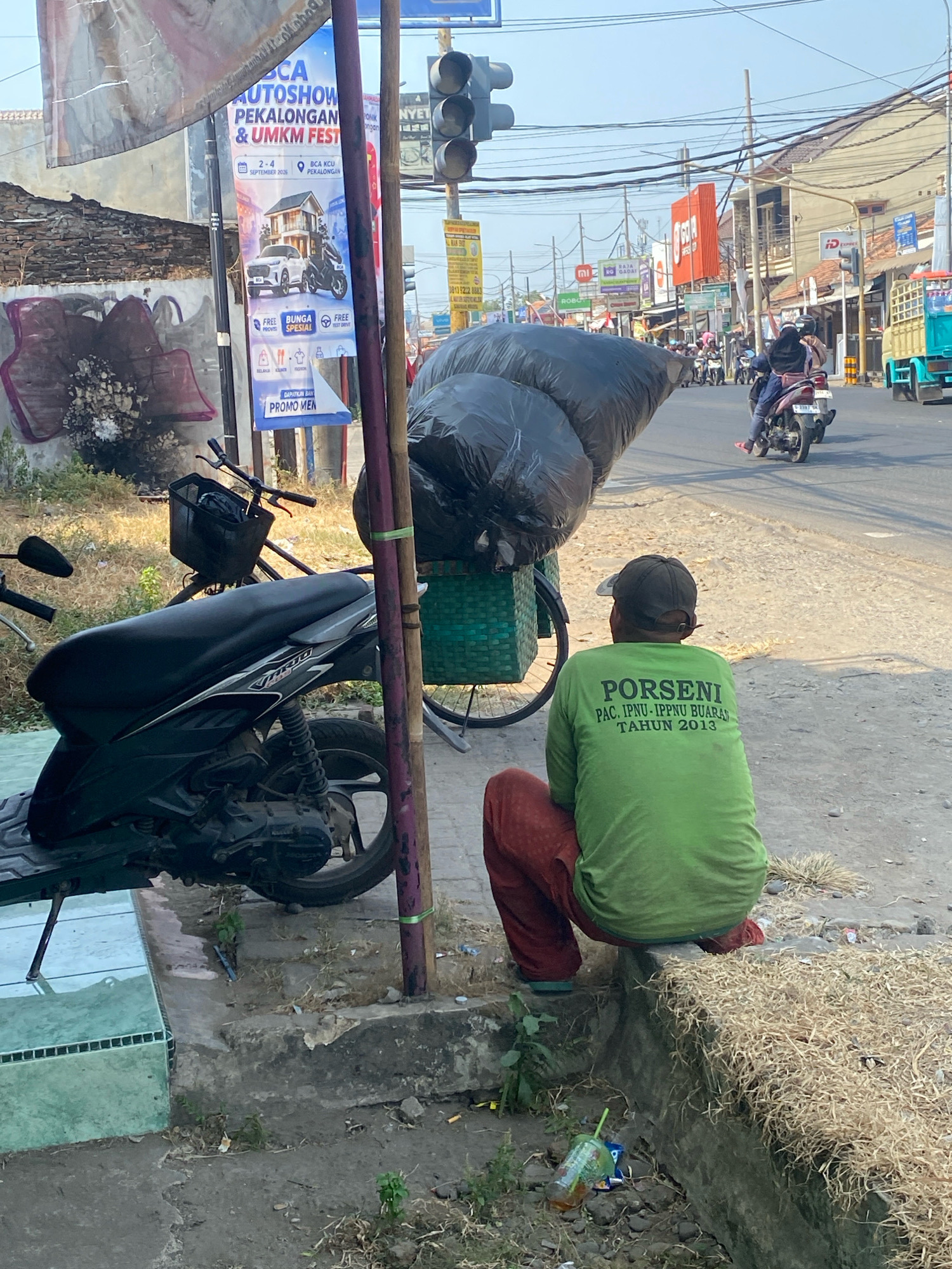The image depicts a street scene in Podo, Indonesia, likely during daylight hours. In the foreground, a person wearing a green long-sleeved shirt with the text "PORSENI PAC. IPNU-IPPNU BUARAN TAHUN 2013" and red pants is squatting on a raised concrete edge. They are facing away from the camera. To the left of this person, a dark-colored motorcycle is parked on a tiled surface. A bicycle with a green basket is positioned behind the motorcycle, carrying two large black garbage bags. In the middle ground, a busy street is visible with several motorcycles and a truck. Traffic lights are present in the background, along with various buildings and storefronts. The overall environment appears to be an urban setting with evidence of everyday activity and some litter on the ground.