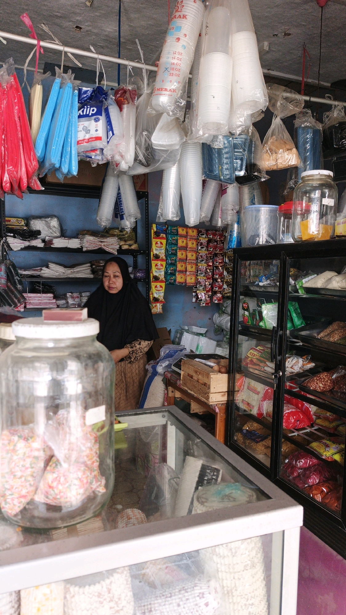 A woman wearing a black hijab and a patterned dress stands behind a counter in a small shop in Cibenda, Indonesia. The shop appears to be a convenience store or a small grocery store. The shelves behind her are stocked with various items, including packaged food, drinks, and disposable cups. In front of her is a large glass jar filled with colorful sprinkles and a glass display case. The overall impression is that of a busy, well-stocked local shop.