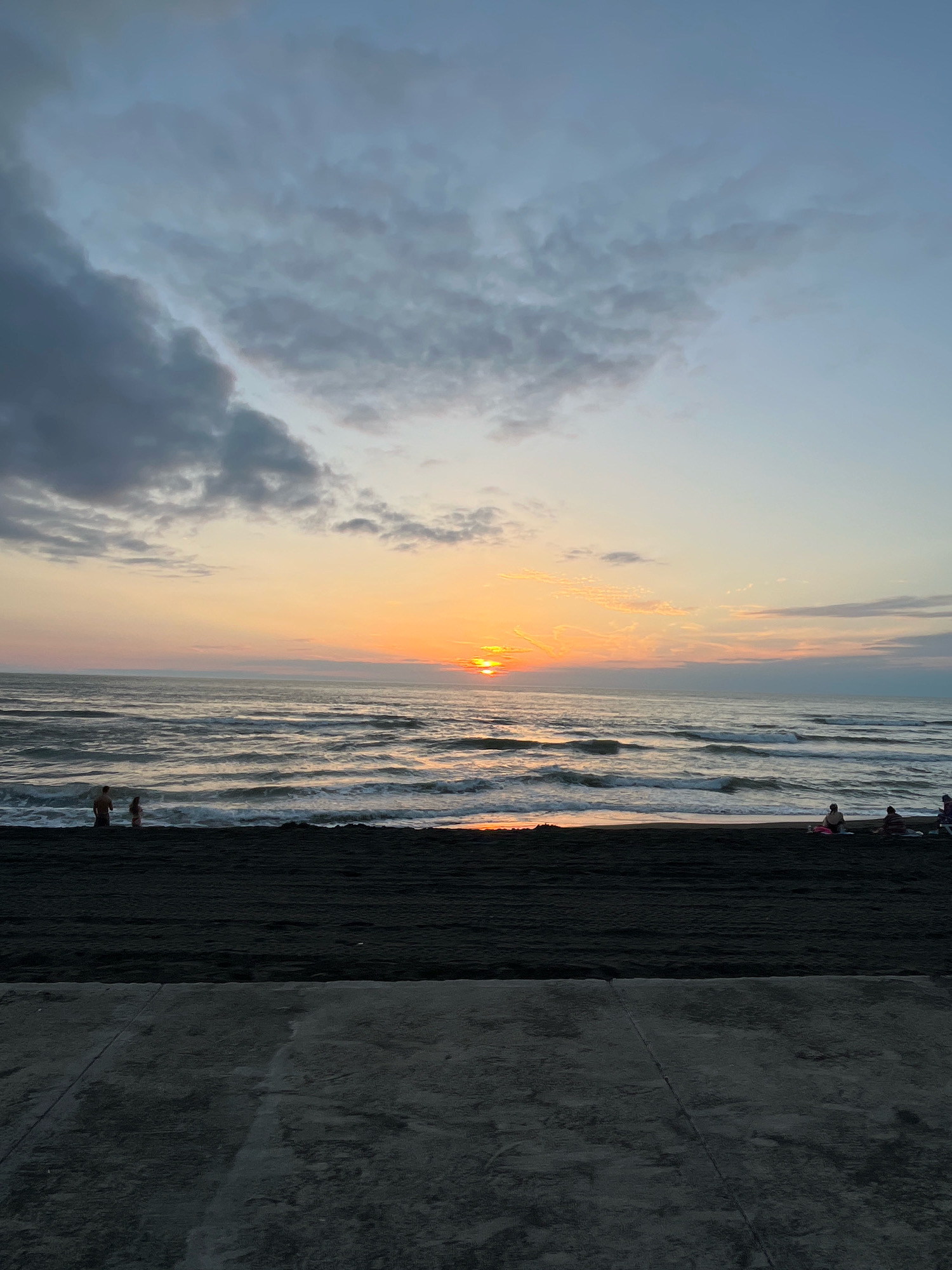 The image depicts a serene beach scene at sunset in Ureki, Georgia. The sun, a bright orange orb, is low on the horizon, casting a warm glow across the sky and reflecting on the wavy ocean. Dark, textured sand forms the foreground and mid-ground of the beach. A few people are visible on the beach: a couple stands near the water's edge, while several others are seated further back, some on towels or loungers. The ocean is a deep blue with white-capped waves rolling towards the shore. The sky above is a gradient of soft blues and oranges, with scattered clouds that are tinged with the sunset's colors. The overall atmosphere is peaceful and tranquil, characteristic of a quiet evening by the sea.