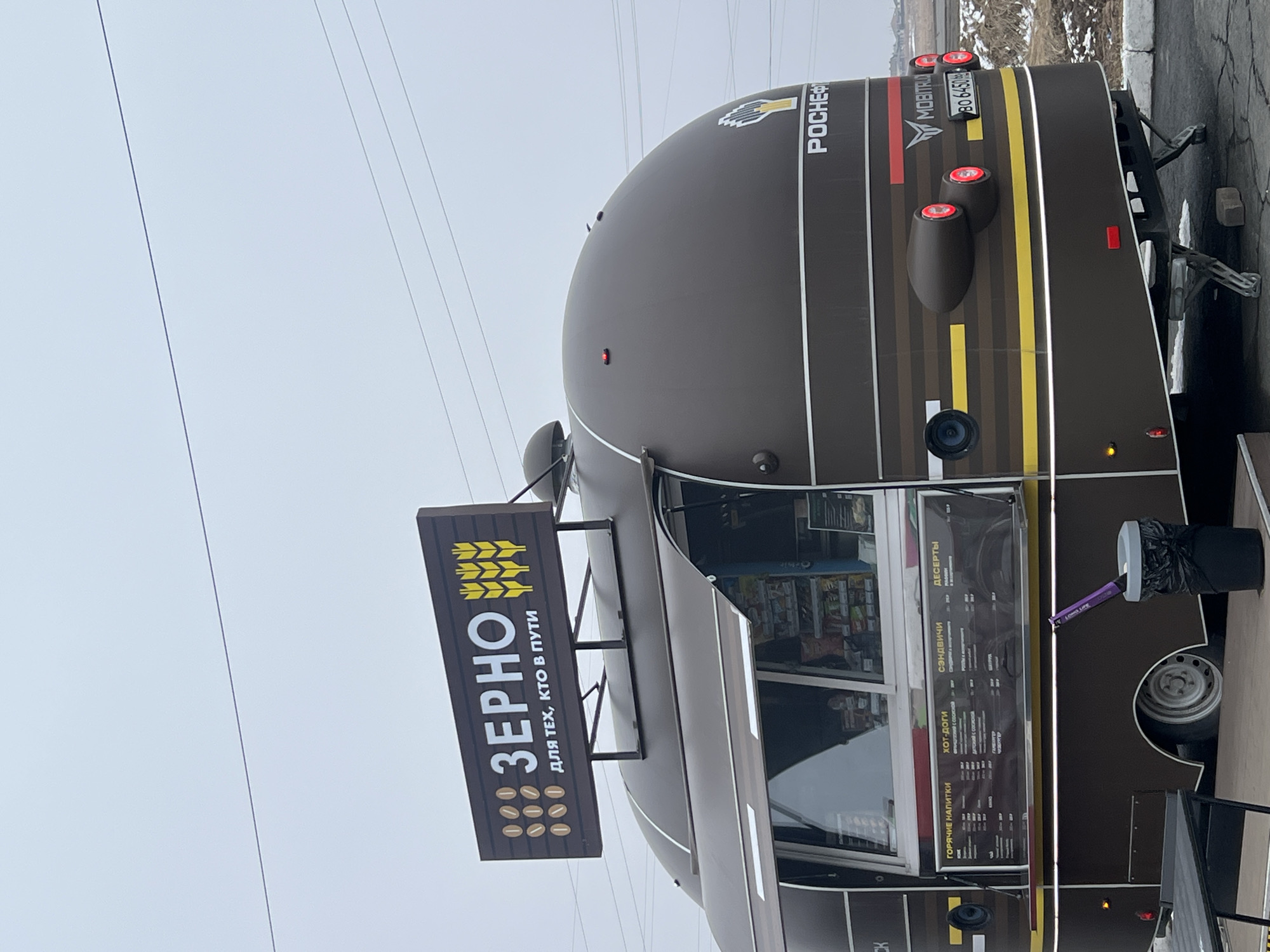 The image depicts a food truck in Kamennoye, Russia. The truck has a brown exterior with a yellow stripe and is branded "POCNEФTb" and "MOBITRUO". A sign reading "ЗЕРНО" (Zerno) is attached to the side, with the phrase "ДЛЯ ТЕХ, КТО В ПУТИ" (For those on the go). The menu visible on the side of the truck lists hot drinks, hot dogs, sandwiches, and desserts. There's a trash can beside the truck. Power lines are visible in the background.