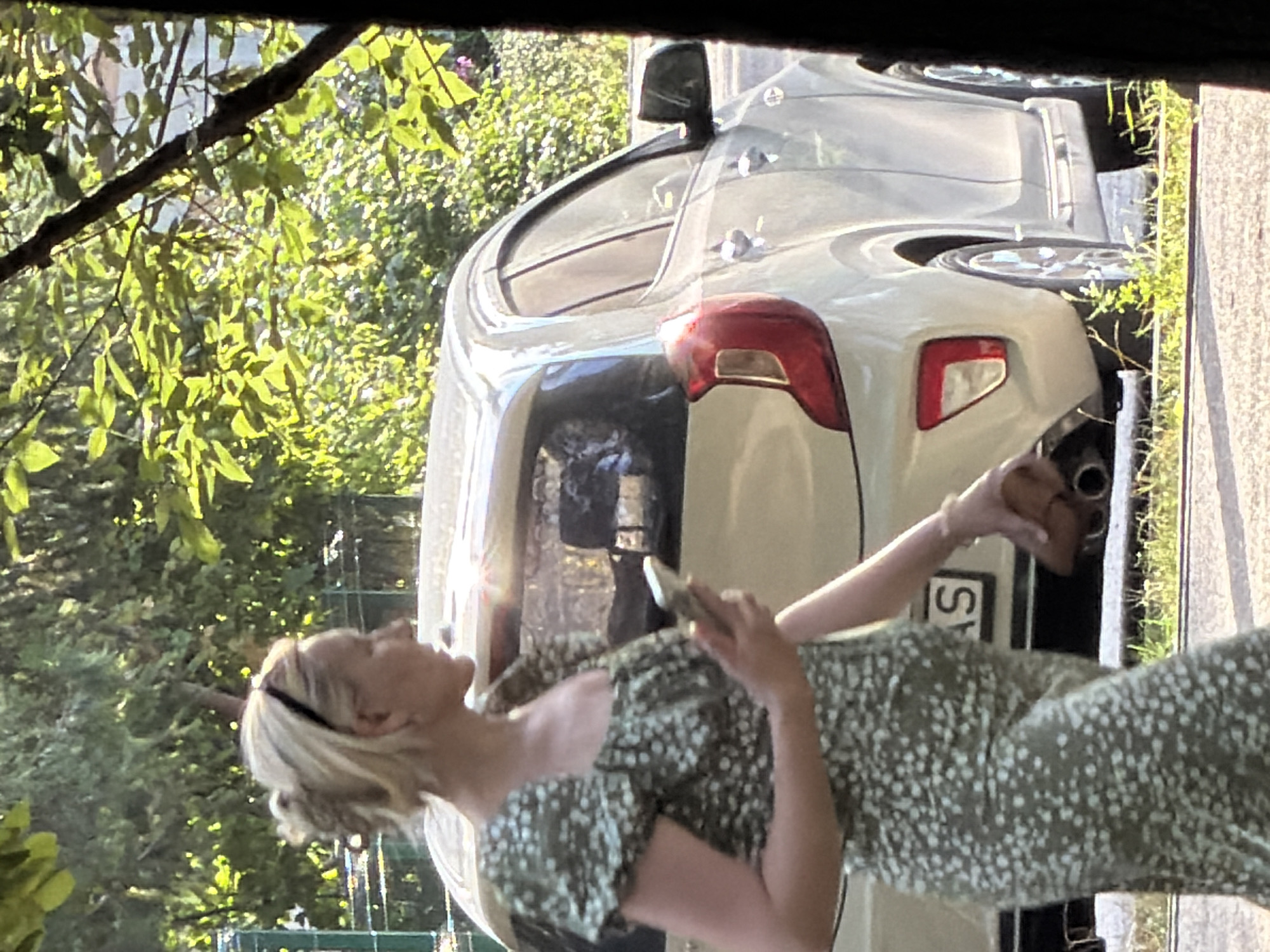 A woman with blonde hair pulled back in a bun is walking and looking at her phone. She is wearing a green floral-print dress with a square neckline. She is holding a brown purse in her right hand. A light-colored car is parked behind her, and trees and foliage are visible in the background. The scene appears to be outdoors on a sunny day.