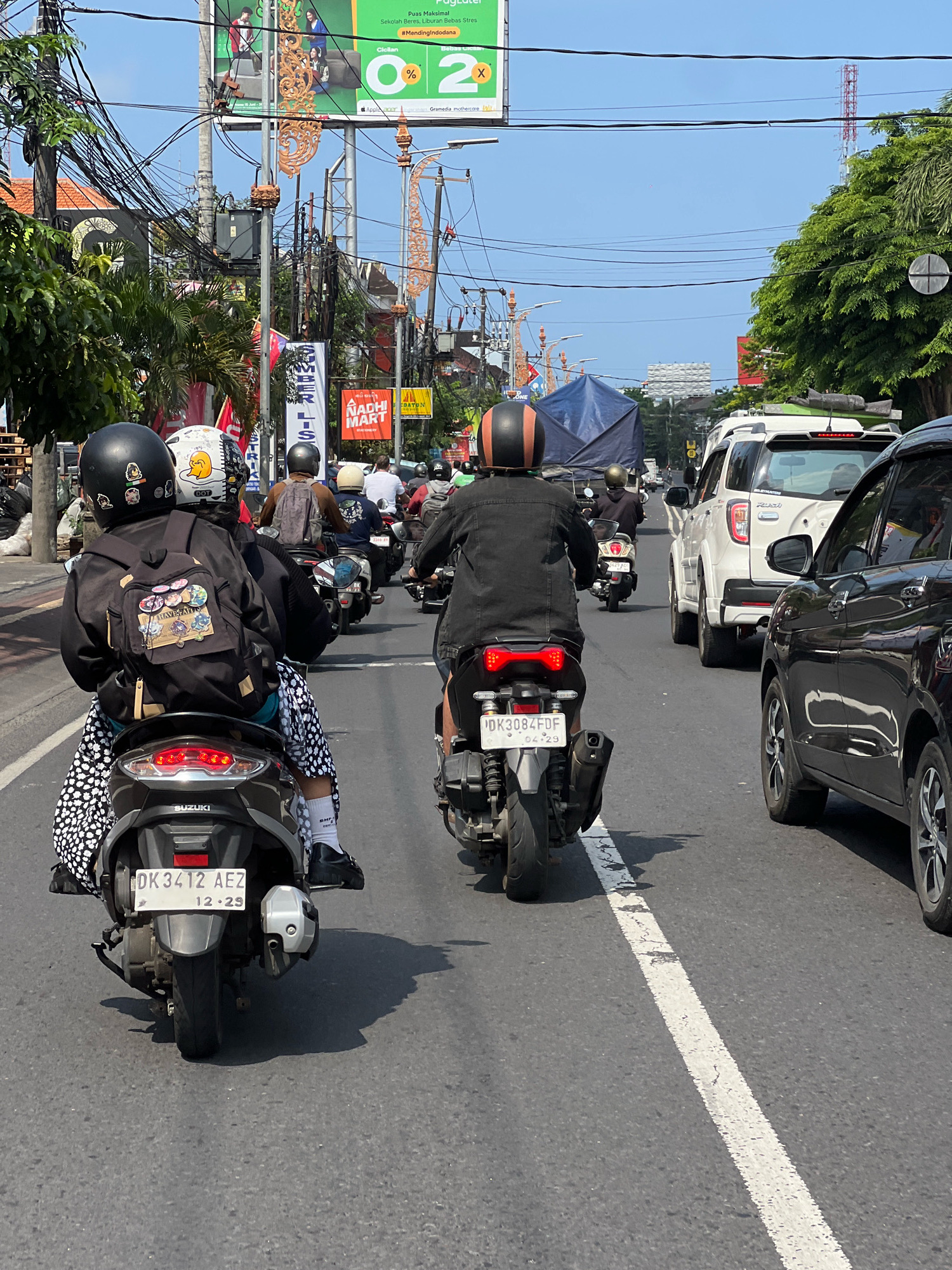 A sunny day in Denpasar, Indonesia, finds a traffic jam on a busy street. The foreground features two motorcycles, one with a black jacket and patterned pants, and another with a black denim jacket. Both riders wear helmets. Behind them, other motorcycles and cars navigate the congestion. Buildings and power lines line the street, with lush green trees visible in the distance. Billboards with advertisements are perched above the street, indicating a commercial area. The road itself is asphalt with a white dividing line, and shadows of the vehicles stretch across it.