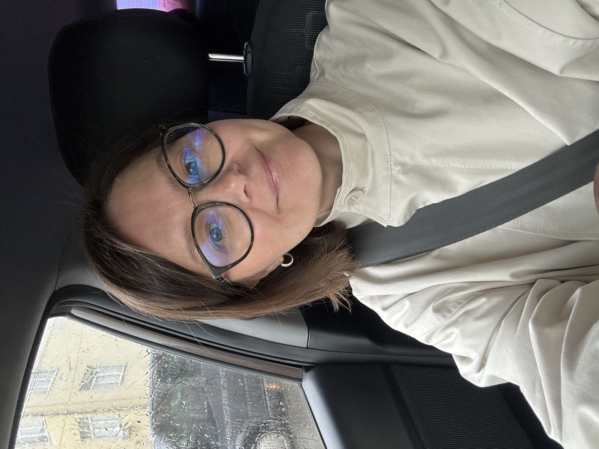 A woman wearing glasses and a light-colored jacket is in a car, buckled up with a seatbelt. The window is covered in raindrops, obscuring the view outside. Based on visual cues such as architectural details and window construction, the location may be inferred as Izhevsk, Russia.