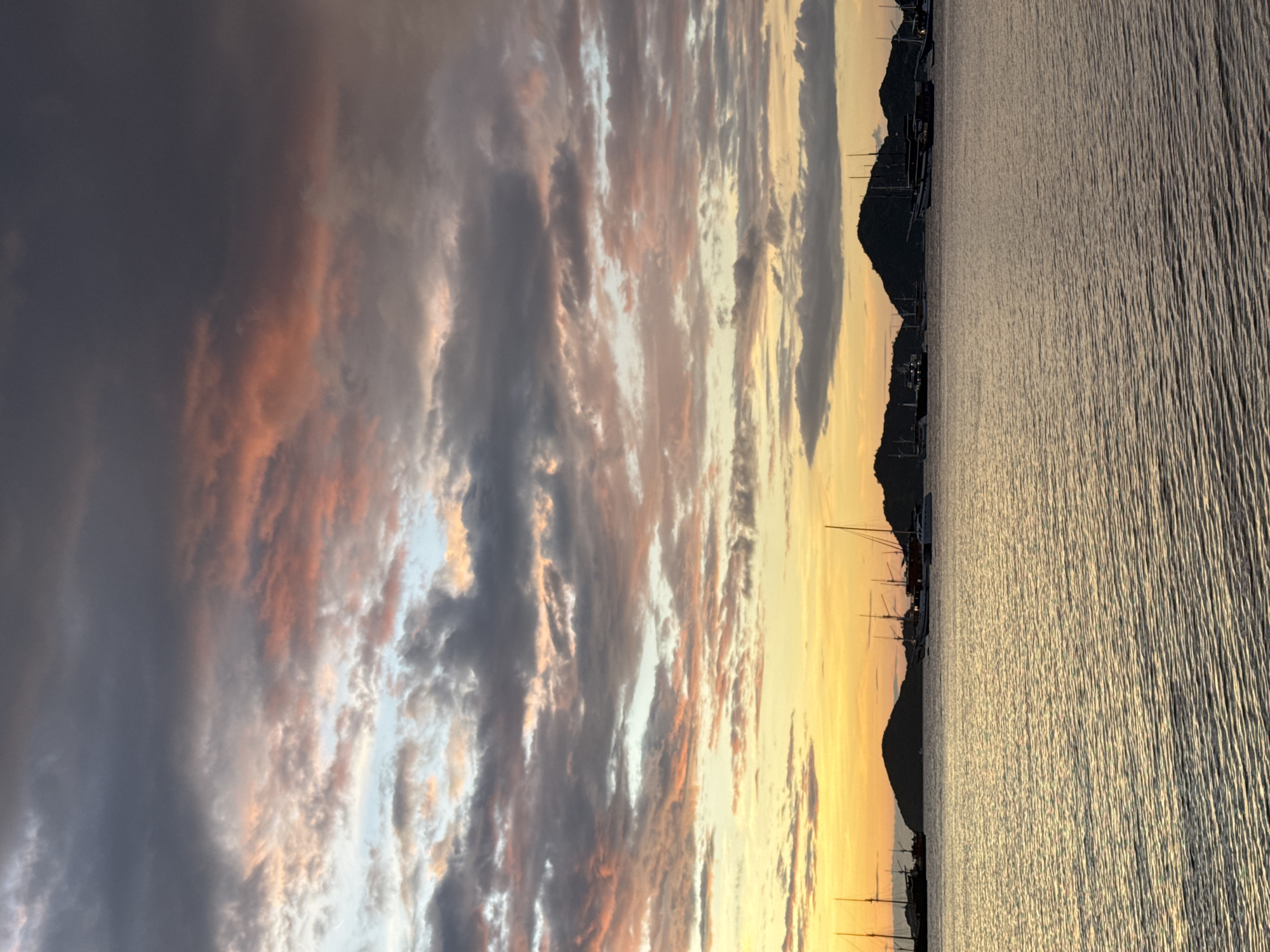 The image shows a sunset over water, likely a harbor, with a silhouetted shoreline and boats. The sky has dramatic clouds with hues of gray, orange, and yellow. The water reflects the sunlight, creating a shimmering effect. The location appears to be a coastal area.