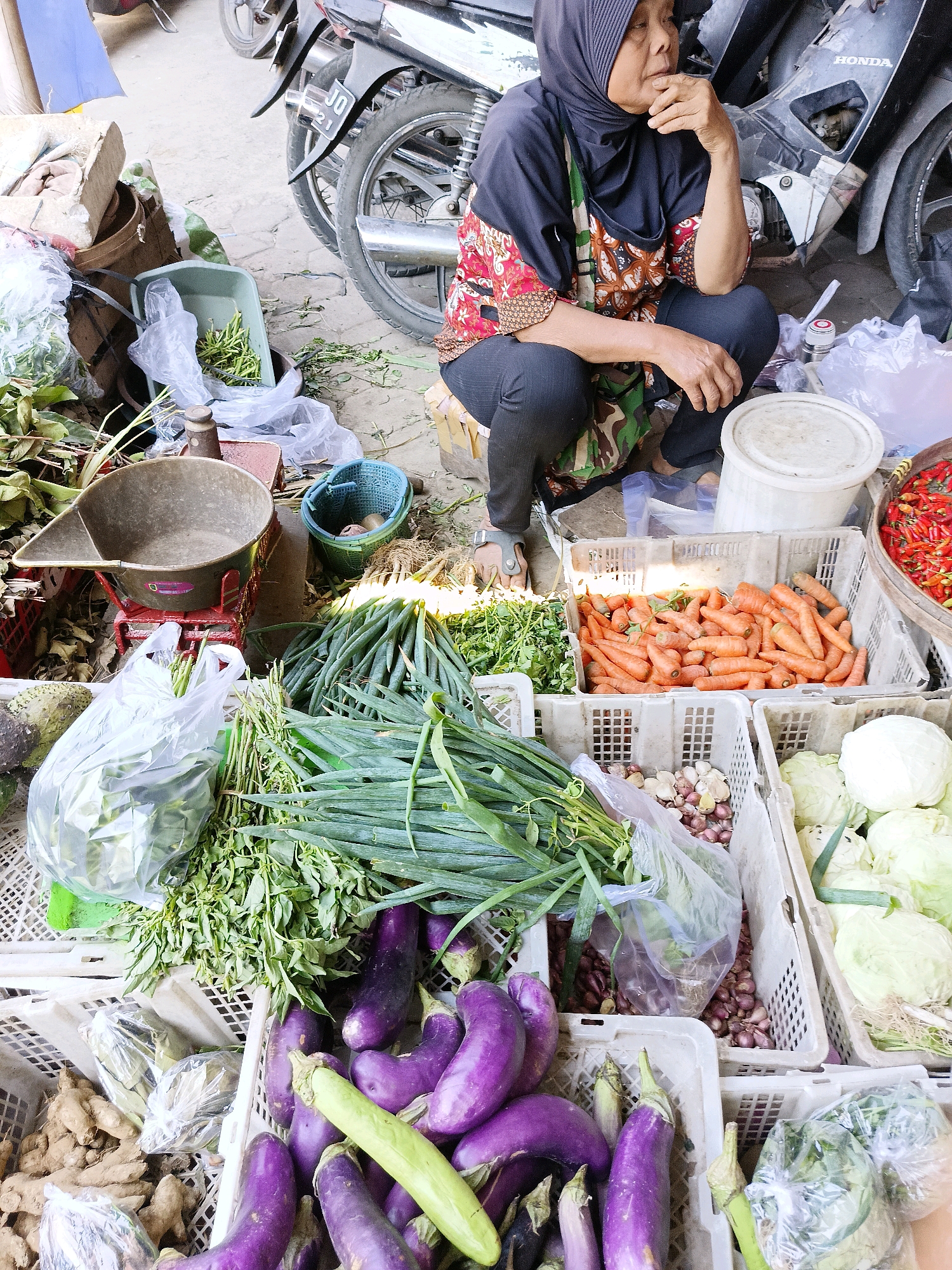 In Manisrenggo, Indonesia, an older woman wearing a hijab and a patterned top is crouching beside a market stall. She has her hand near her mouth, in a pensive gesture. In front of her, various fresh vegetables are displayed in plastic crates. These include bundles of green onions, carrots, eggplants, cabbage, and shallots. To the left of the crates, a traditional scale with a large metal pan is visible, along with other produce in plastic bags and baskets. A motorcycle is parked behind the woman, partially obscuring the background. The ground appears to be concrete or dirt, typical of an outdoor market setting.
